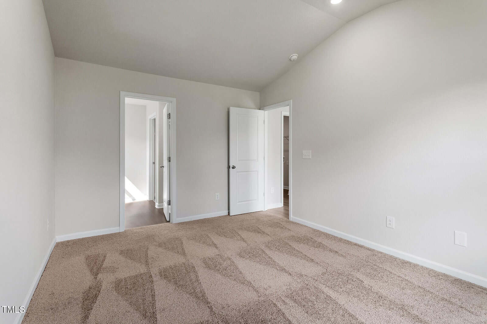 An empty room with light beige walls and carpeted floor. The room features two open doors; one leads to another room with natural light, and the other to a corridor. The ceiling is vaulted and there are no furnishings.