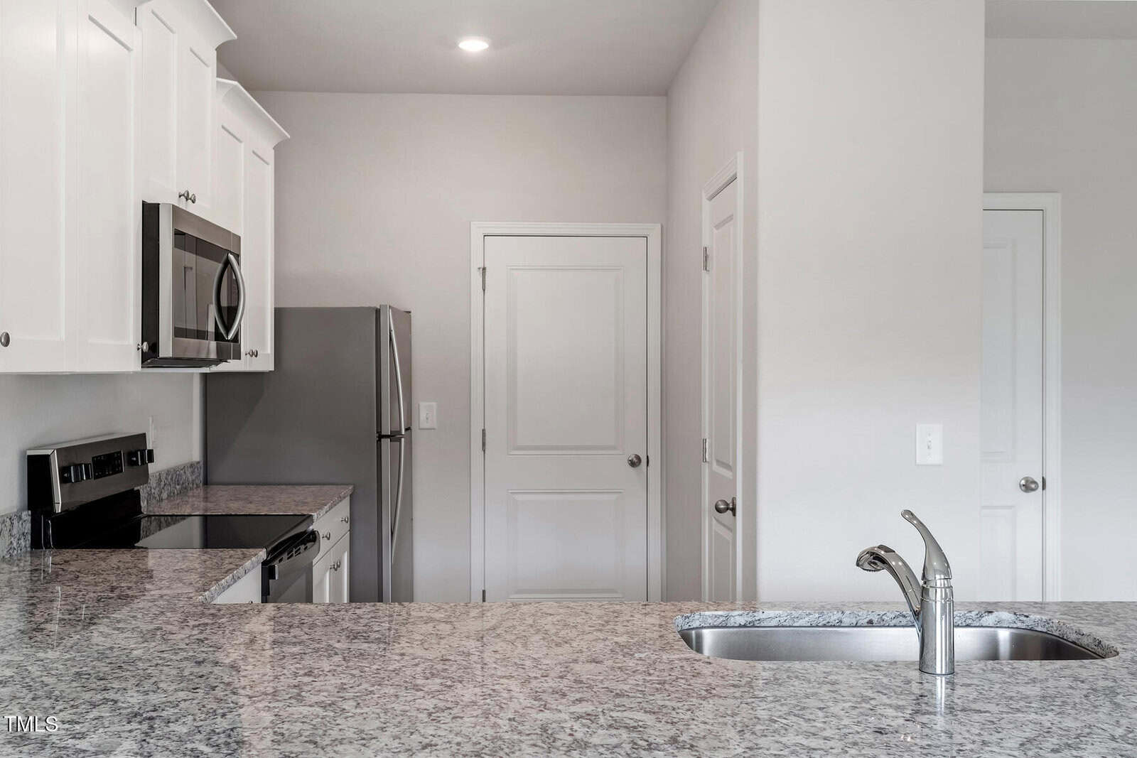 A modern kitchen with white cabinets, stainless steel appliances, and a granite countertop. The scene includes an electric stove, microwave, refrigerator, and a sink with a faucet on the island. Two closed white doors are situated in the background.
