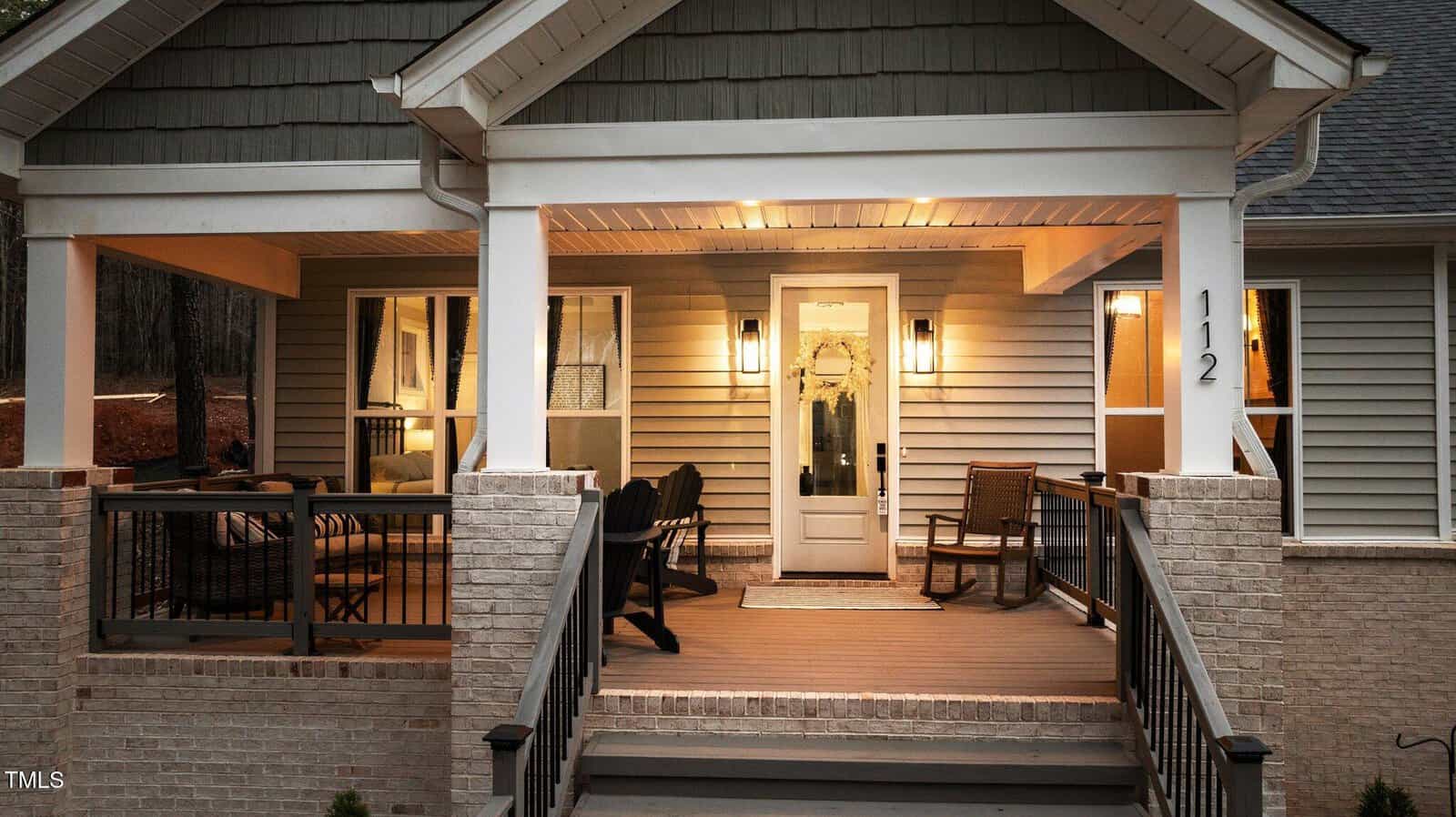 A well-lit house front with a covered porch, white pillars, beige siding, black railing, a glass front door, rocking chairs, and patio furniture. The house number 112 is visible near the door. Trees appear in the background.