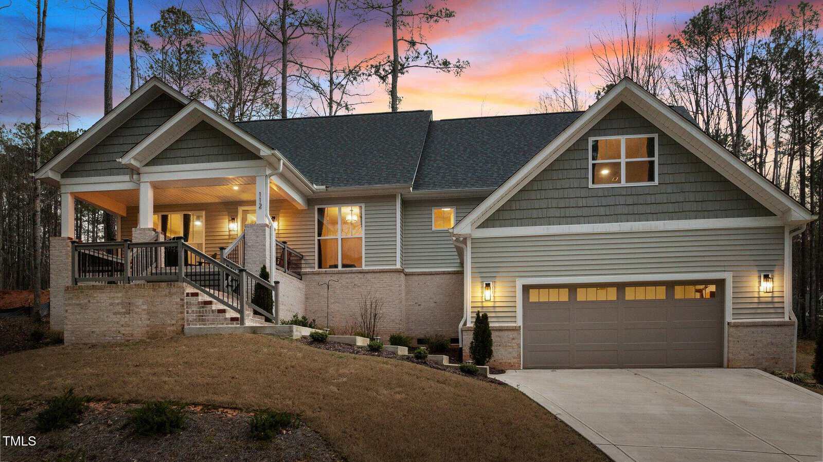Single-story house at 112 Rawhide Drive, Louisburg, featuring a welcoming front porch, double garage, brick and siding exterior, sloped driveway, and lit windows at dusk—all surrounded by trees beneath a partly cloudy sky tinged with sunset colors in the background.