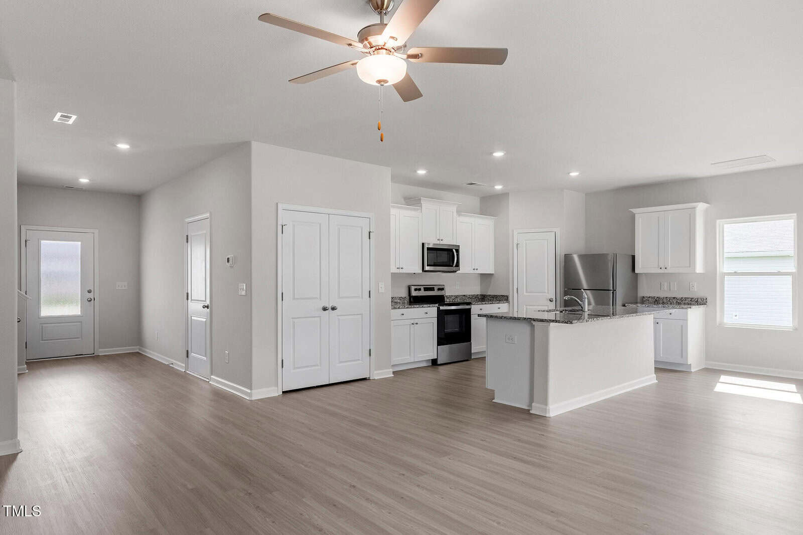 A modern kitchen and living area with light wood flooring. The kitchen features white cabinets, stainless steel appliances, and a center island with a granite countertop. A ceiling fan is mounted in the living space. Brightly lit with natural light.