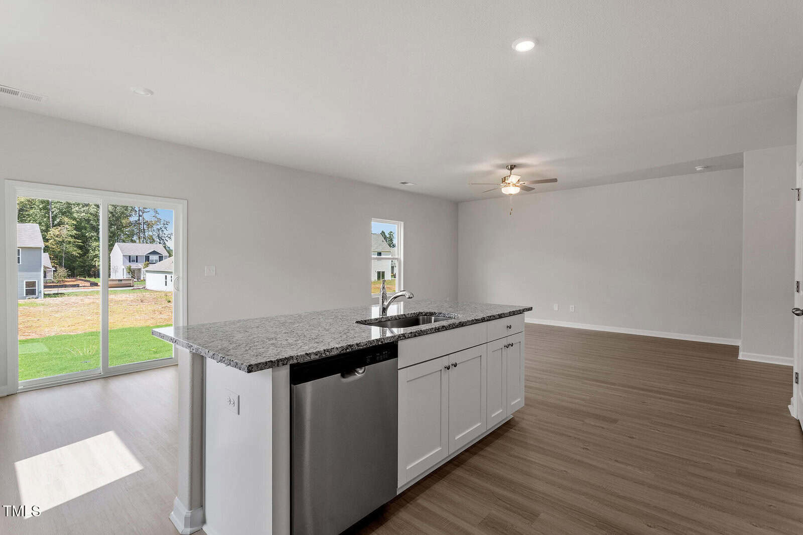 A spacious, empty modern kitchen with light wood flooring. The kitchen features a gray granite countertop island with a built-in sink and dishwasher. In the background, there's a large living area with a ceiling fan and a sliding door leading outside.
