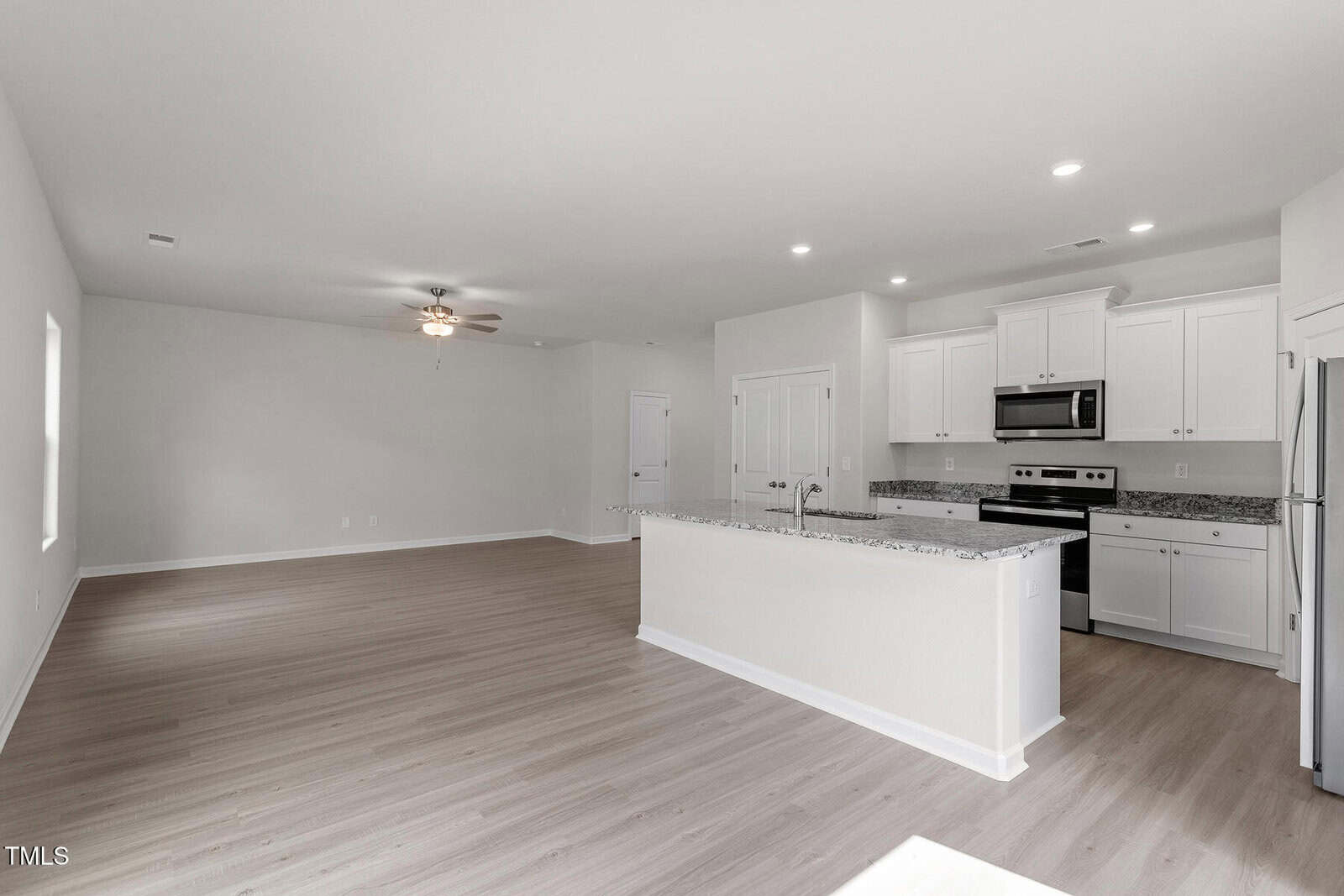 A modern, unfurnished open-concept kitchen and living area. Kitchen features white cabinets, granite countertops, stainless steel appliances, and an island with a sink. Living area has wood flooring, ceiling fan, and recessed lighting.