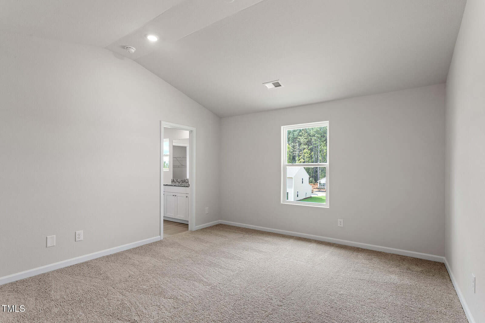An empty room with beige carpet and light gray walls. A single window on the right wall lets in natural light. An open doorway on the left leads to another room with a view of a kitchen. The ceiling is vaulted with recessed lighting.