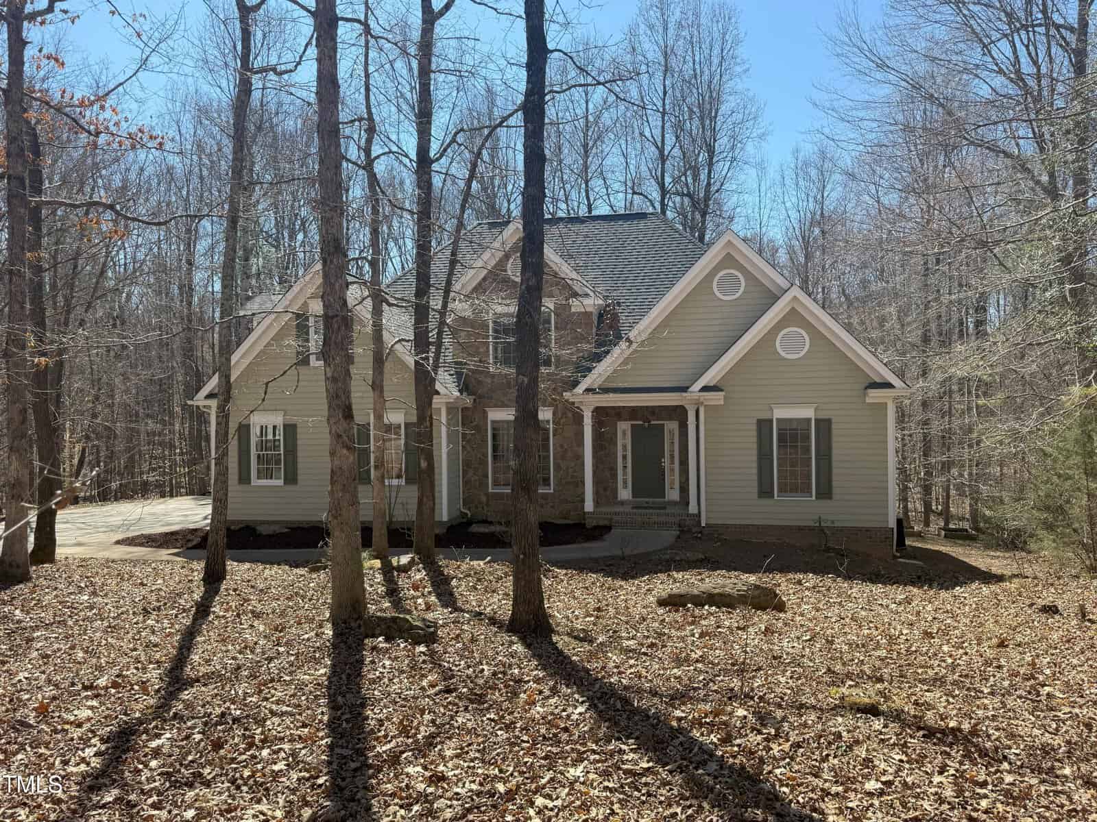 A two-story house with beige siding, brick accents, and a dark gray roof stands among tall, leafless trees. The front yard is covered with fallen leaves, and a driveway is visible on the left side.