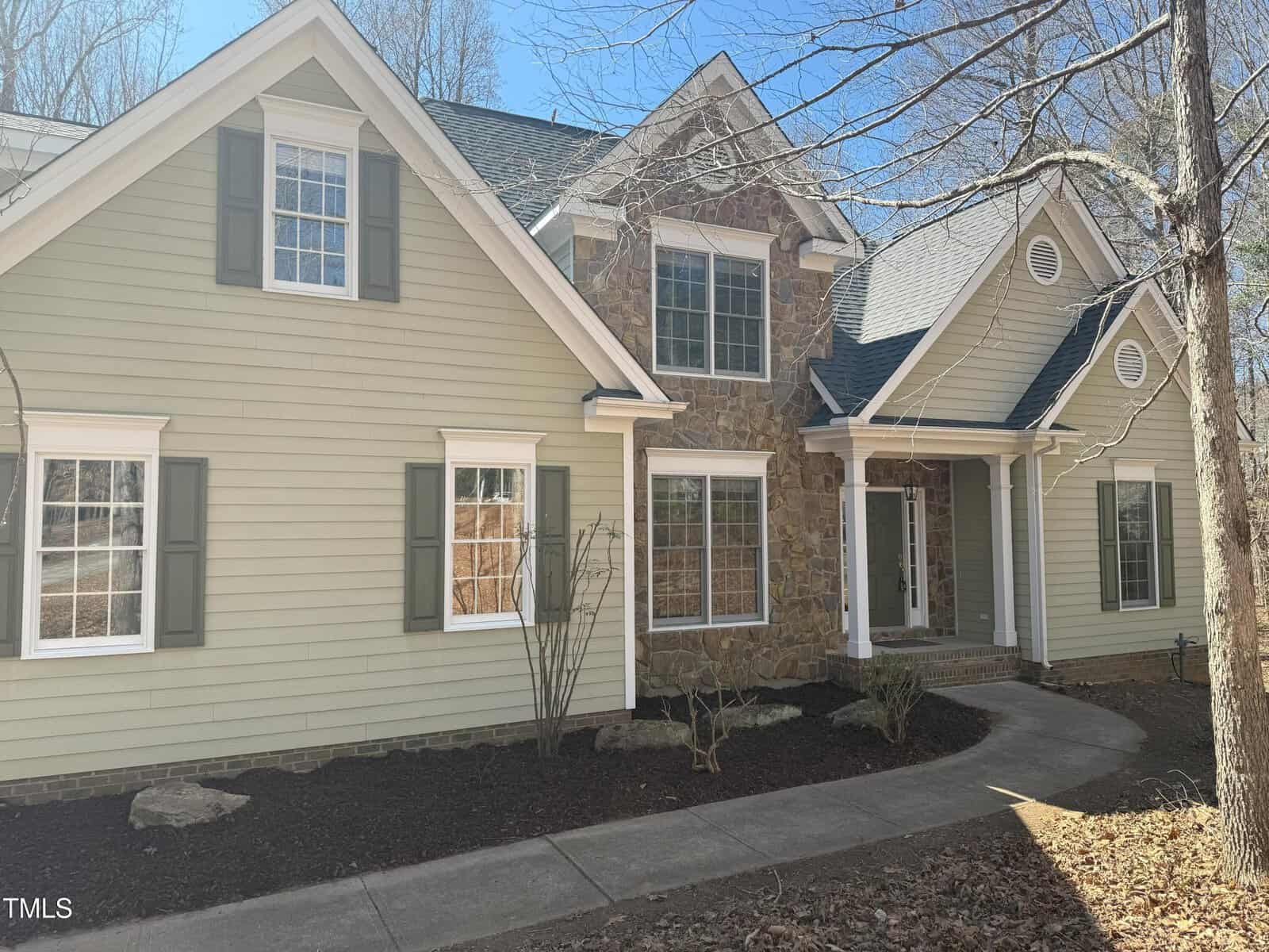 A two-story house with light green siding, stone accents, white trim, and gray shutters. There are several windows, a covered entryway with columns, bare landscaping, and a concrete walkway in front.