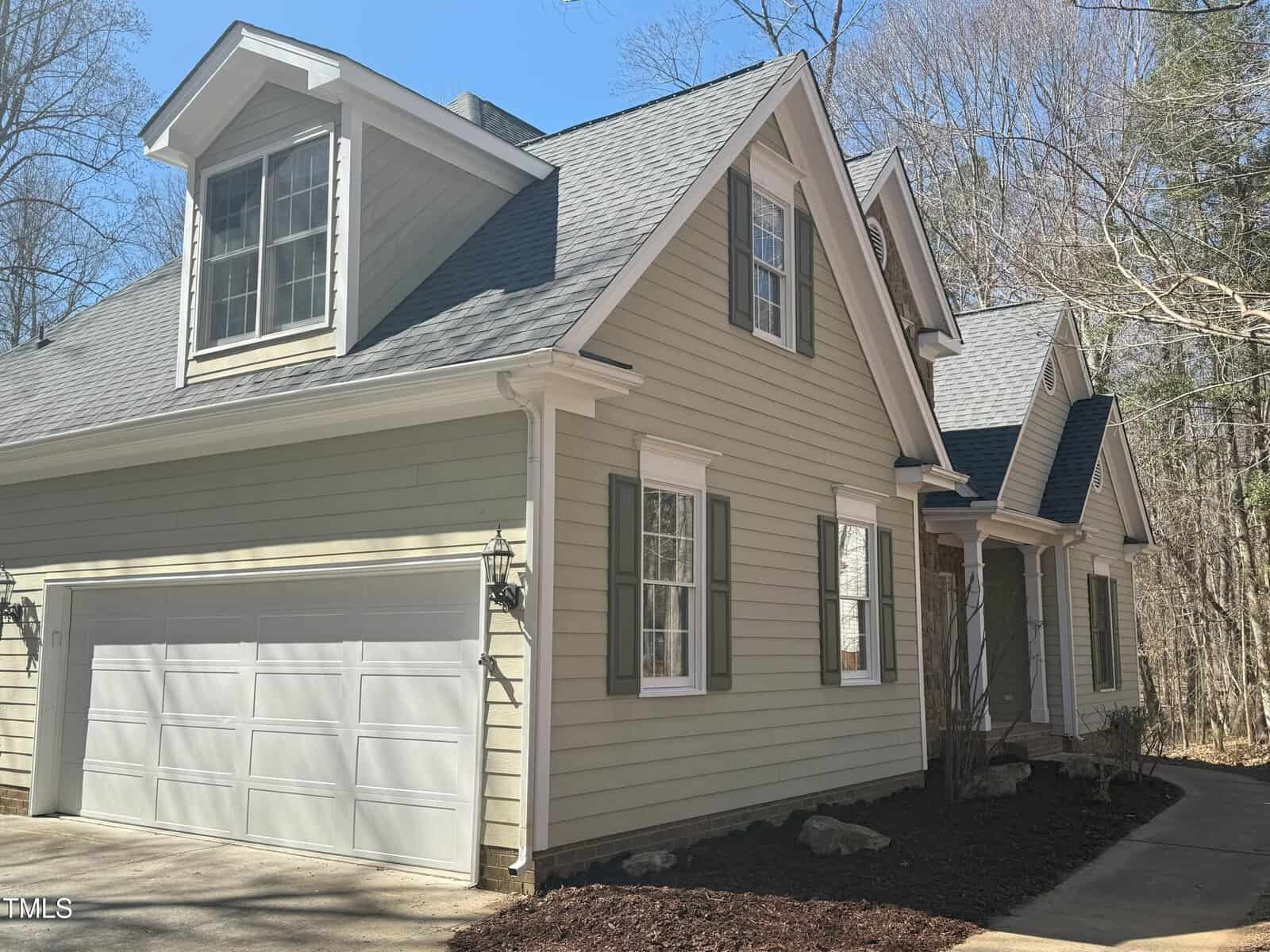 A light gray two-story house with white trim, a double garage, and dormer windows. The house has green shutters, a paved driveway, and is surrounded by bare trees and a mulched garden area.