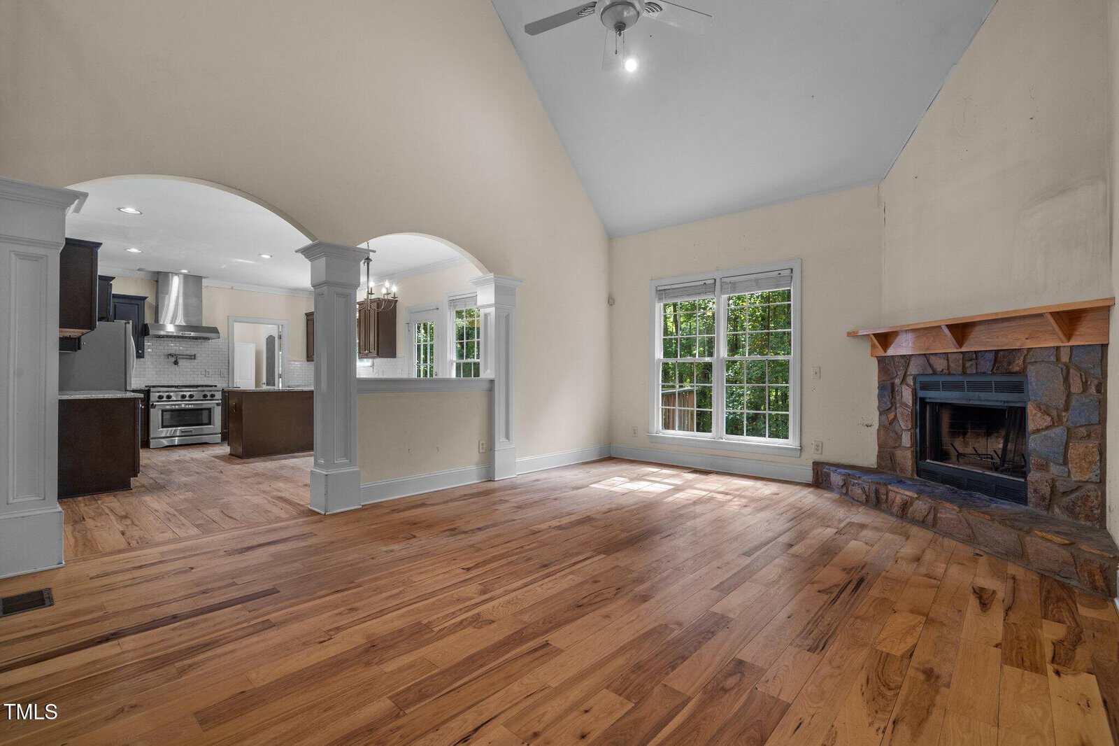 Spacious living area with hardwood floors, a stone fireplace, large windows, and an open view into the kitchen with stainless steel appliances, arched openings, and natural light.