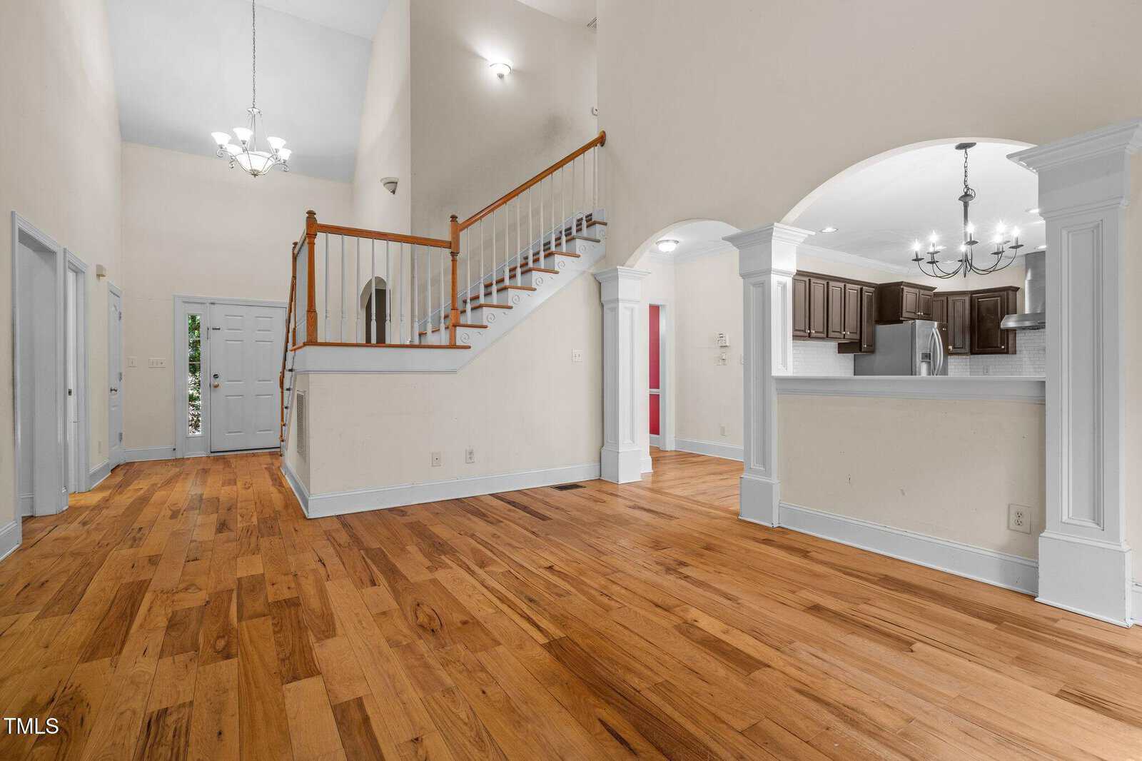 Interior of a home with hardwood floors, white walls, a staircase with wooden railing, an open kitchen with dark cabinets, and an arched opening. Multiple doors and chandeliers are visible. The space is empty and well-lit.