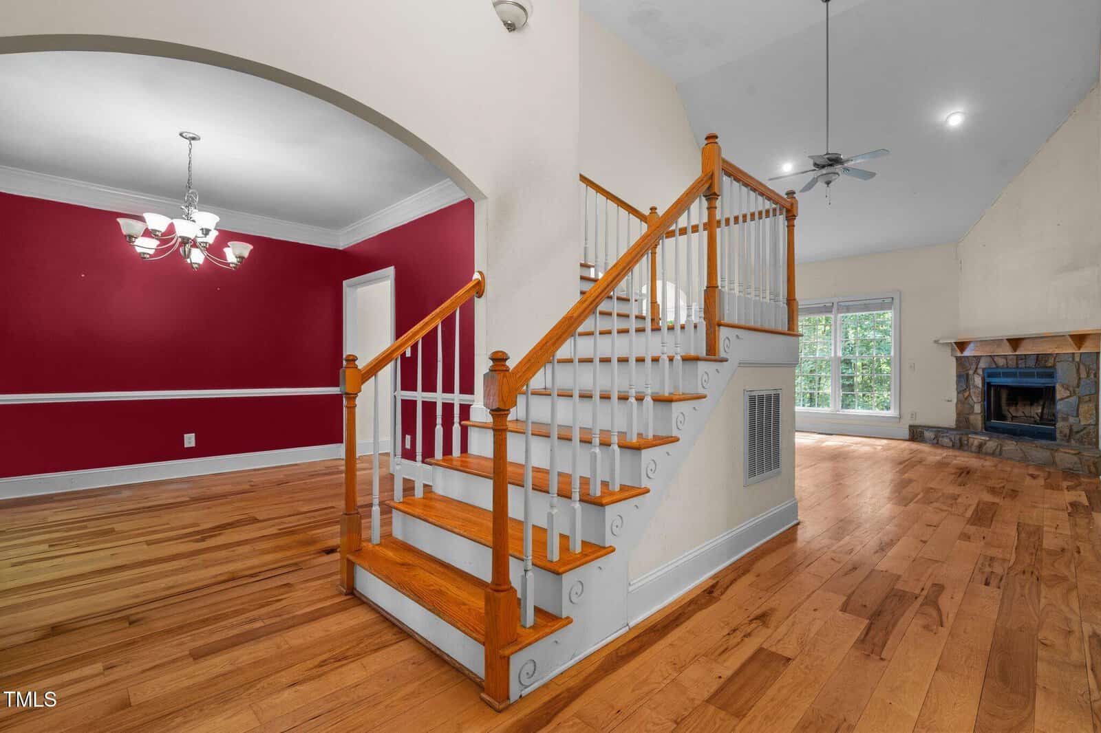 A wooden staircase with white railings is in the center of a room with hardwood floors. To the left is a dining area with red walls and a chandelier. The living area has a stone fireplace and sliding glass doors.