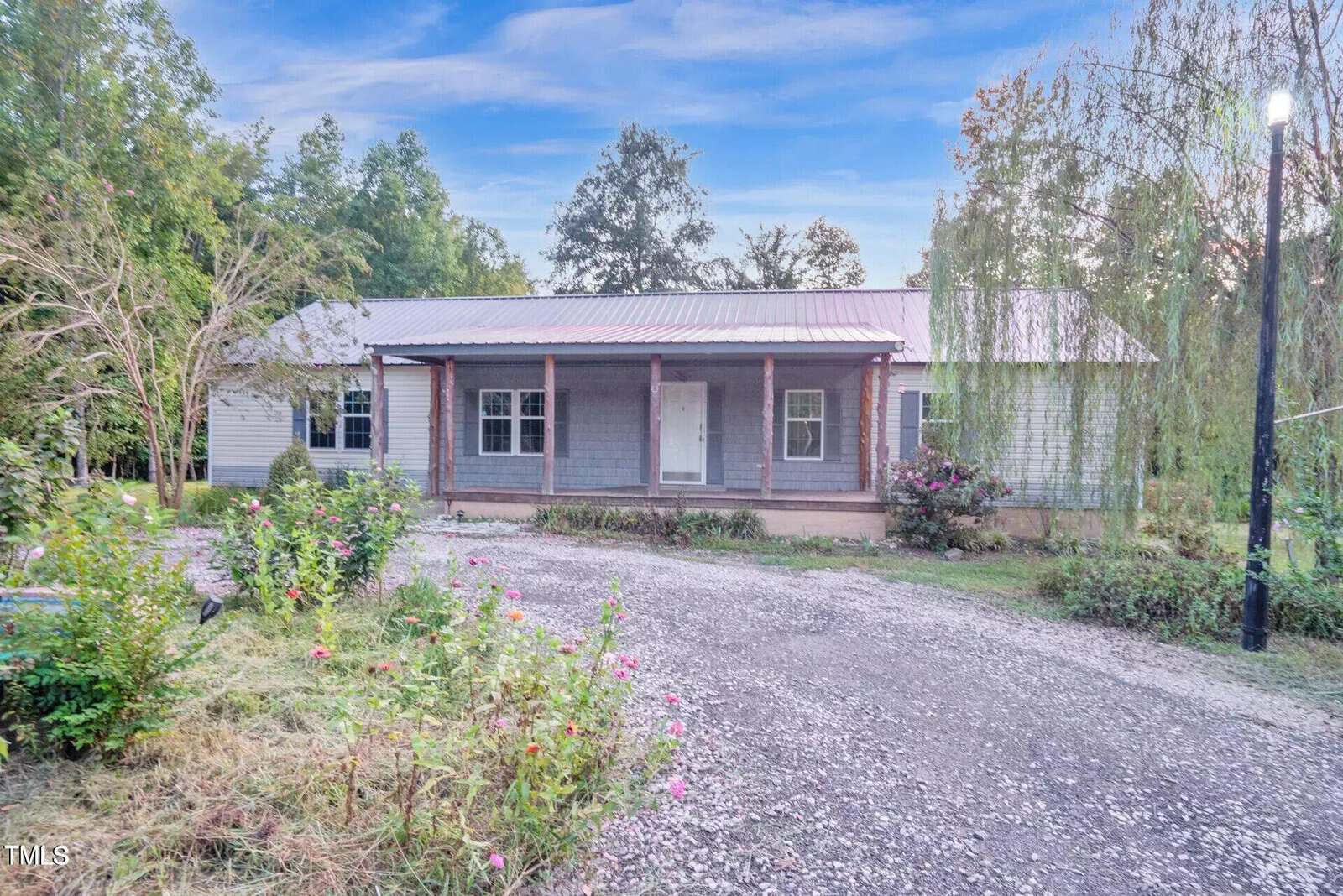 Located at 80 Twin Ponds Lane, this charming single-story Bullock home features a metal roof and a welcoming covered porch. Surrounded by lush trees and shrubs, the gravel driveway is lined with vibrant flowering plants. A lamppost stands to the right as clouds drift across the sky.