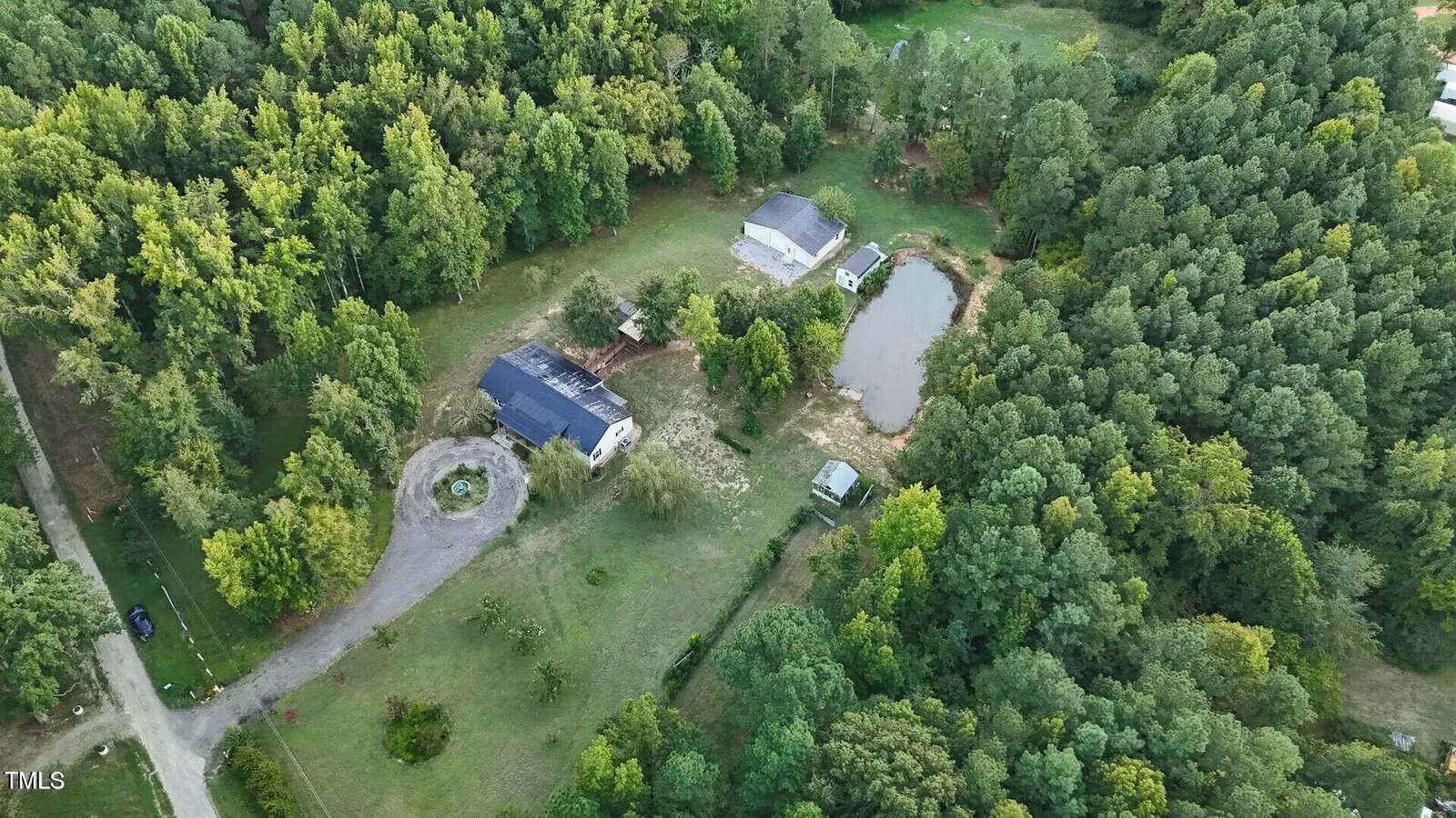 Aerial view of a rural landscape featuring a circular driveway leading to a house surrounded by trees. Adjacent are a small pond and two additional structures. Dense forests border the property on multiple sides.