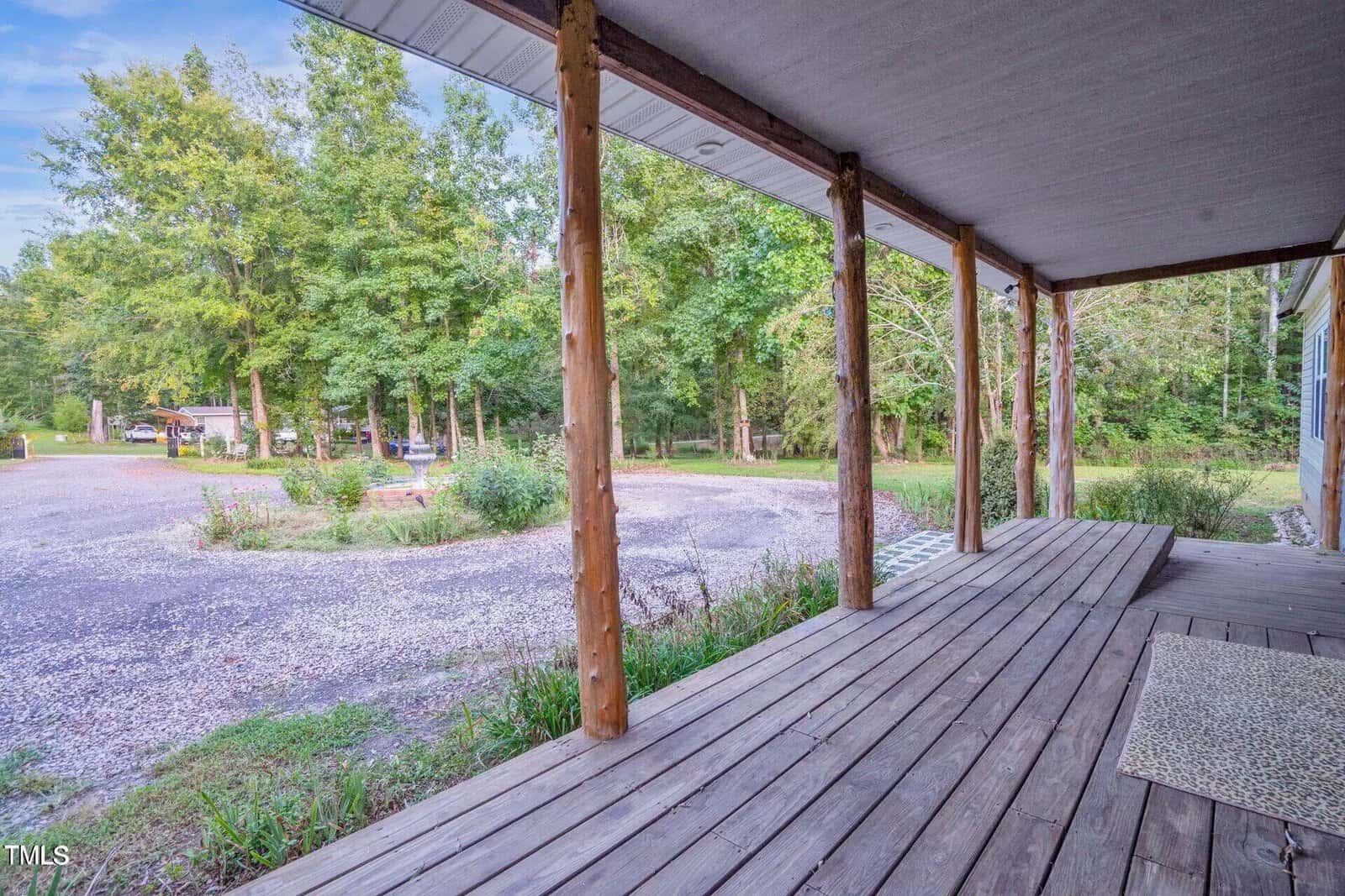 A wooden porch with a view of a gravel driveway and green trees in the background. The porch has wooden beams supporting a roof, and a doormat is placed on the wooden floor. The scene is outdoors, surrounded by nature.