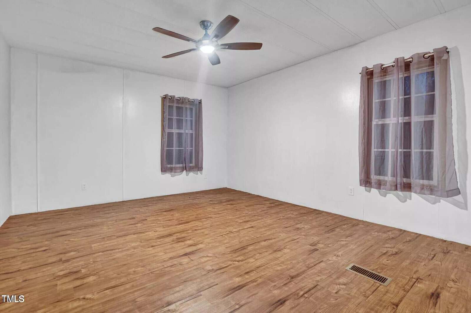 A vacant room with white walls and wooden flooring. It features two windows with sheer brown curtains and a ceiling fan with a light. The room has a simple, clean appearance.