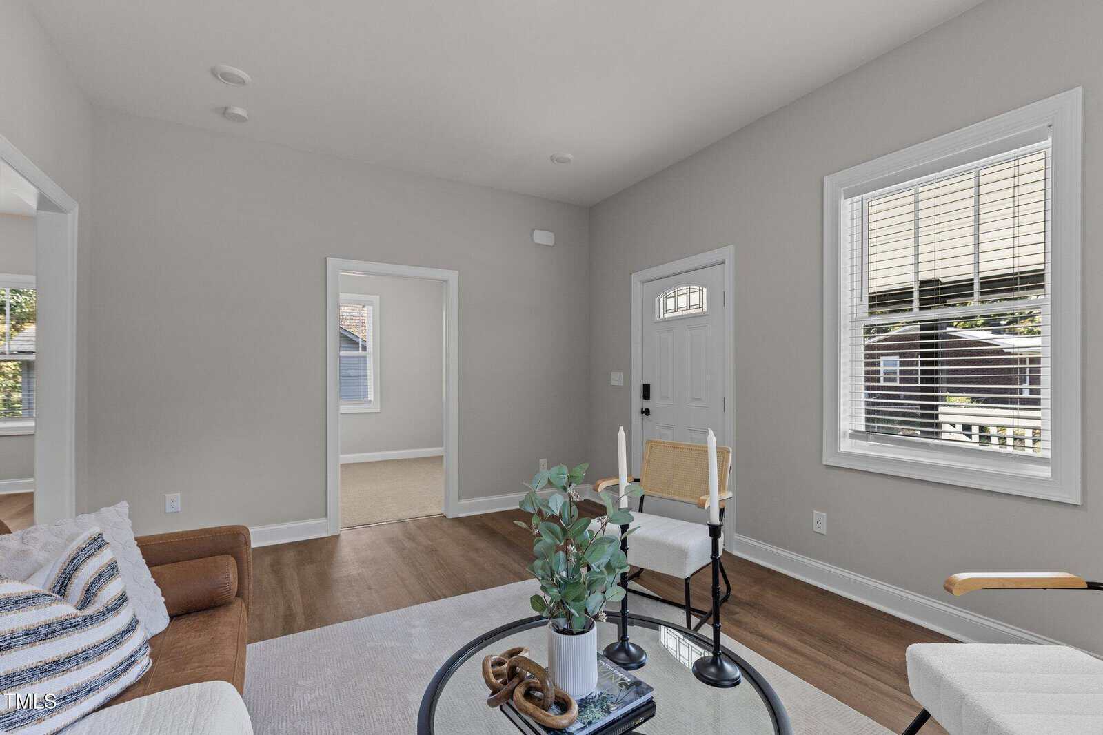 A bright living room with light gray walls and wooden flooring. It features a brown couch with striped cushions, a small round table with plants, a ladder-back chair, and a white front door with windows. A doorway leads to another room.