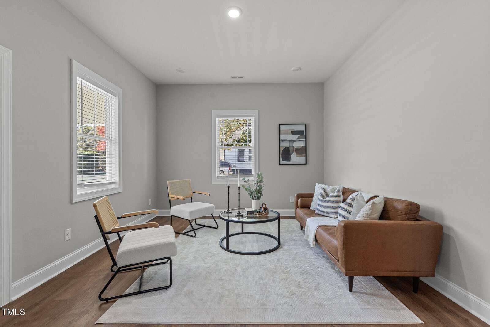 A living room with a brown leather sofa, two white upholstered chairs, a round glass coffee table, and a light gray area rug. Three decorative cushions rest on the sofa. Two windows and a framed picture are on the walls.