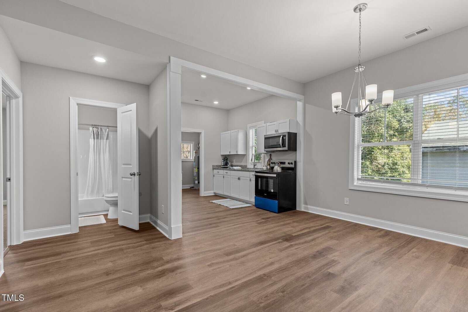 Open concept kitchen and dining area with light grey walls and wood flooring. The kitchen features white cabinets, stainless steel appliances, and a window. Adjacent is a small bathroom with a shower. A chandelier hangs above the dining space.