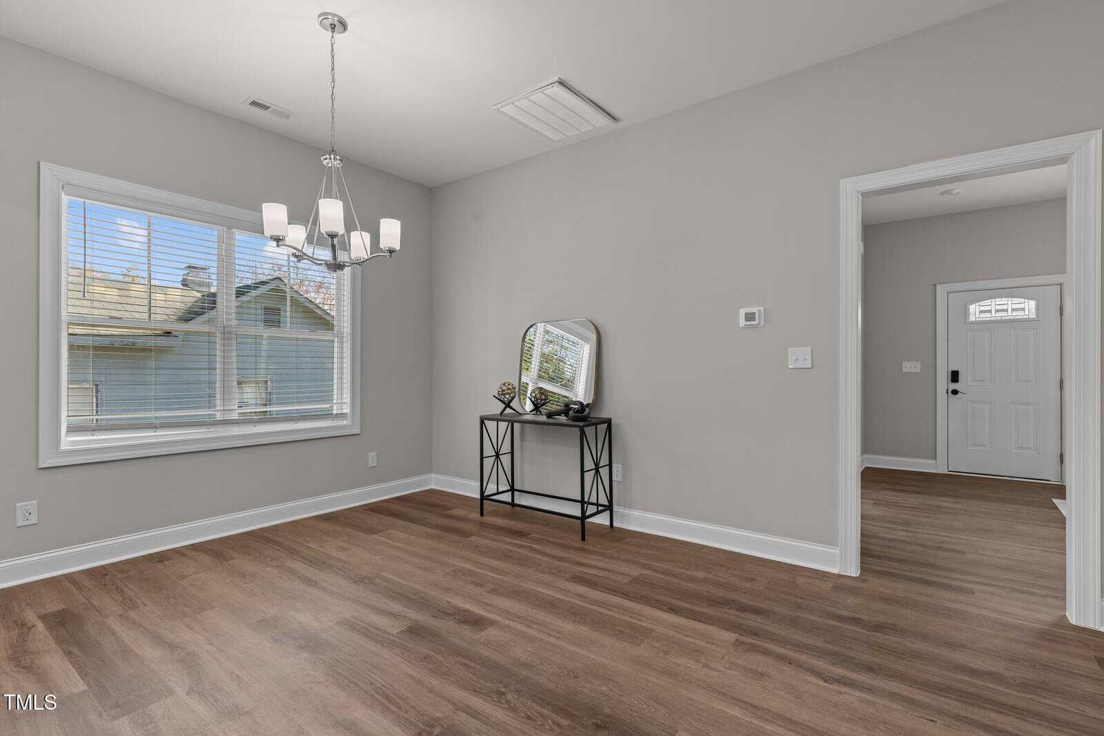 A room with light gray walls and wood flooring, featuring a window with blinds, a ceiling light fixture, and a small table with decor items beneath a wall-mounted mirror. An open doorway leads to another area with a white door.