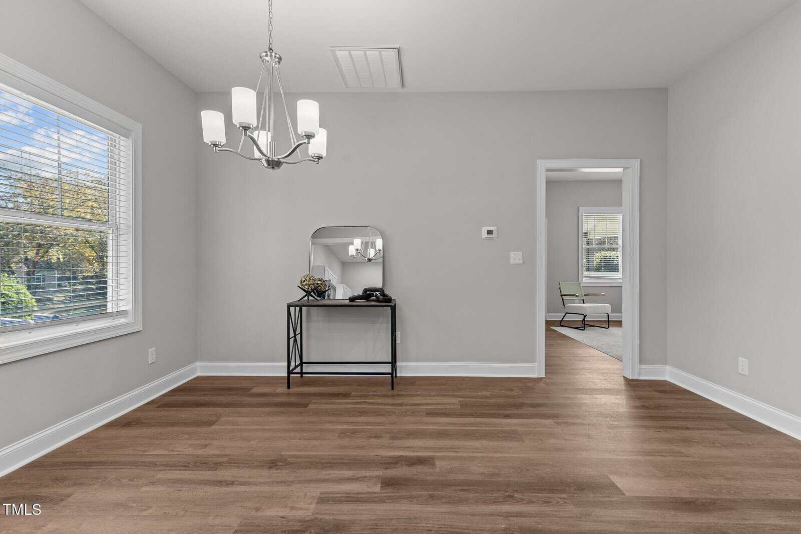 An empty room with light gray walls and wood flooring. A six-light chandelier hangs from the ceiling. A small table with a mirror and a decorative object is against the wall. A doorway leads to another room with a chair and window.