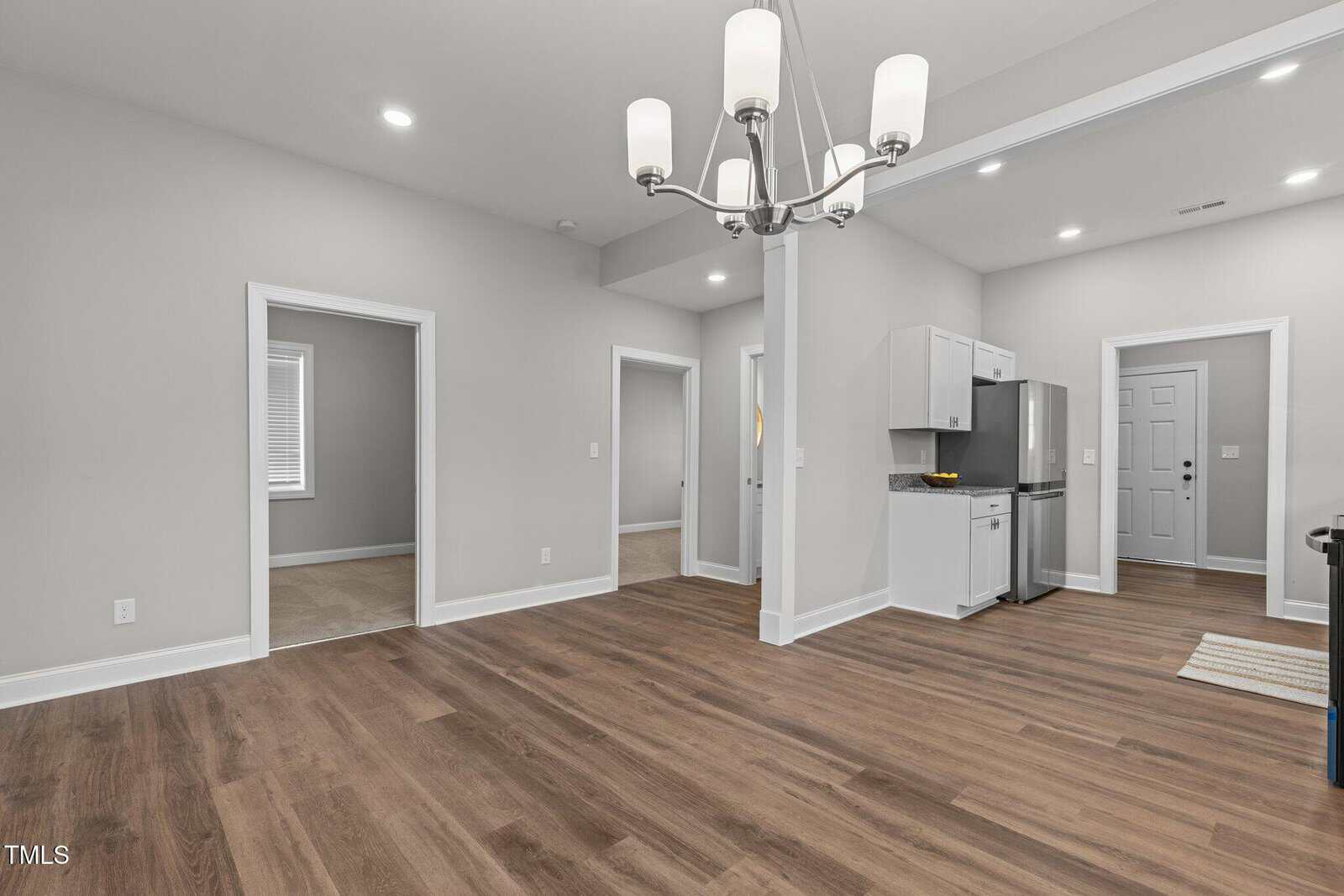 Empty room with hardwood floors, beige walls, and recessed lighting. A chandelier hangs from the ceiling. A small kitchen area with white cabinets and a gray countertop is visible, along with several doorways leading to other rooms.