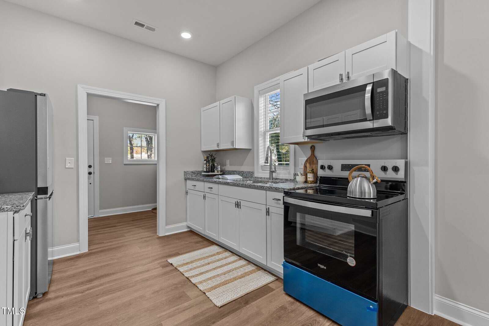A modern kitchen features white cabinets, a stainless steel oven with a kettle on top, and a microwave above. There's a window over the sink, a small indoor plant, and a striped rug on the wooden floor. A door leads to another room.