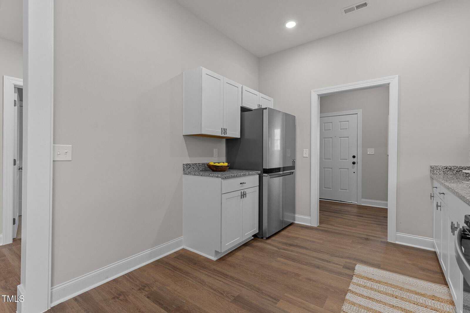 A modern kitchen with light gray walls, white cabinets, stainless steel refrigerator, granite countertops, a fruit bowl, and wood flooring. There is a door and an open doorway visible in the background.