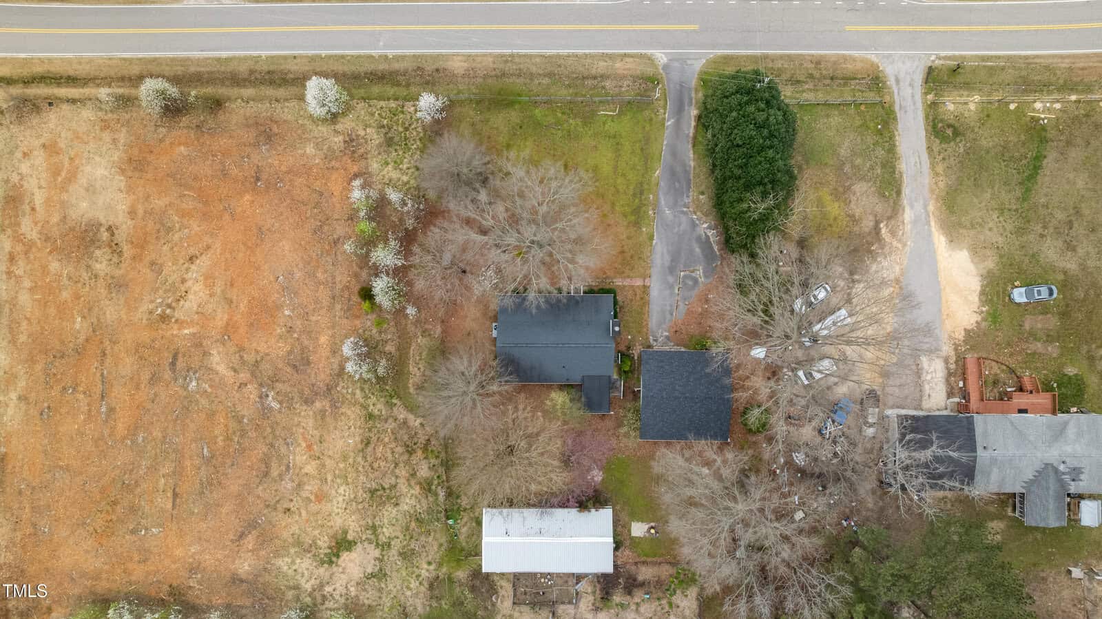 Aerial view of a house with a black roof surrounded by bare trees, a paved driveway with parked cars, adjacent buildings, and a nearby road at the top of the image. Orange-brown and green grass are visible.