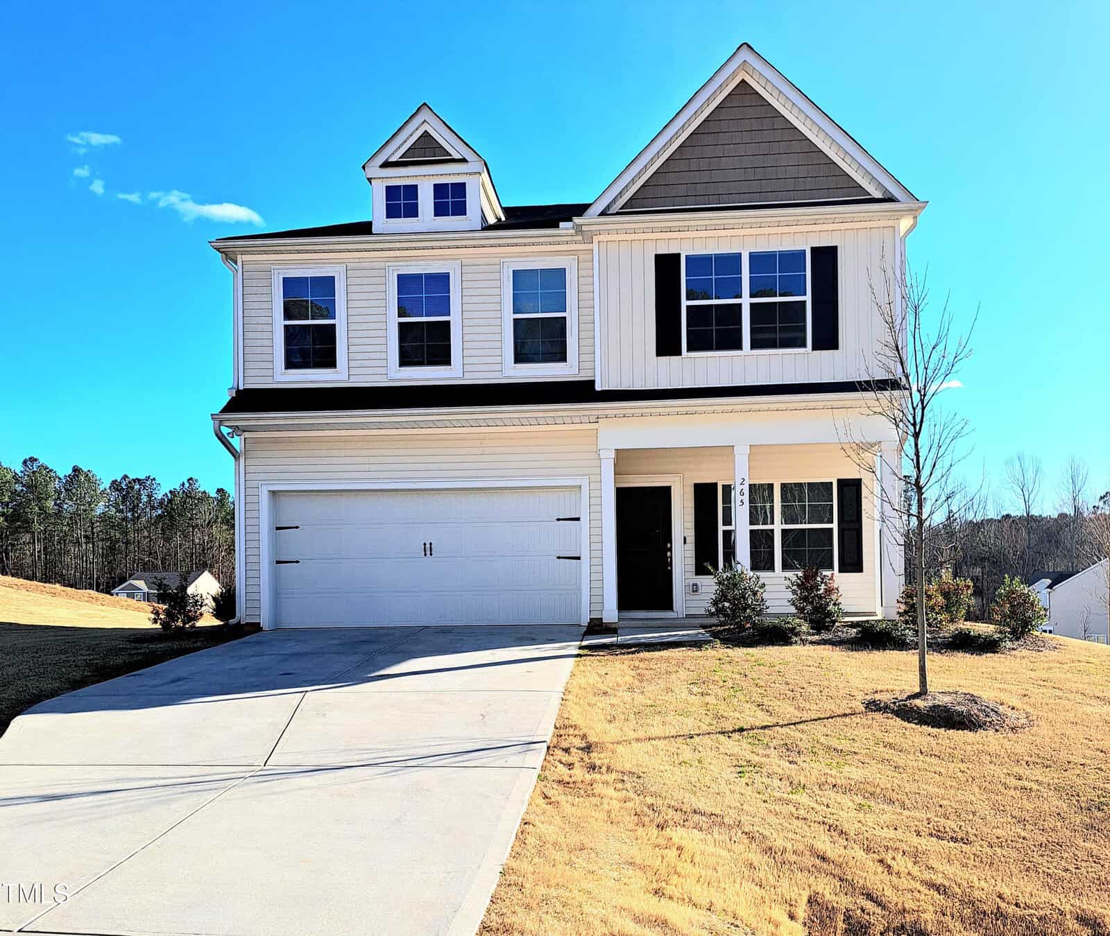 A two-story suburban house with white siding, a two-car garage, black shutters, several front windows, and a small front yard with a tree and bushes, on a sloped lot under a clear blue sky.