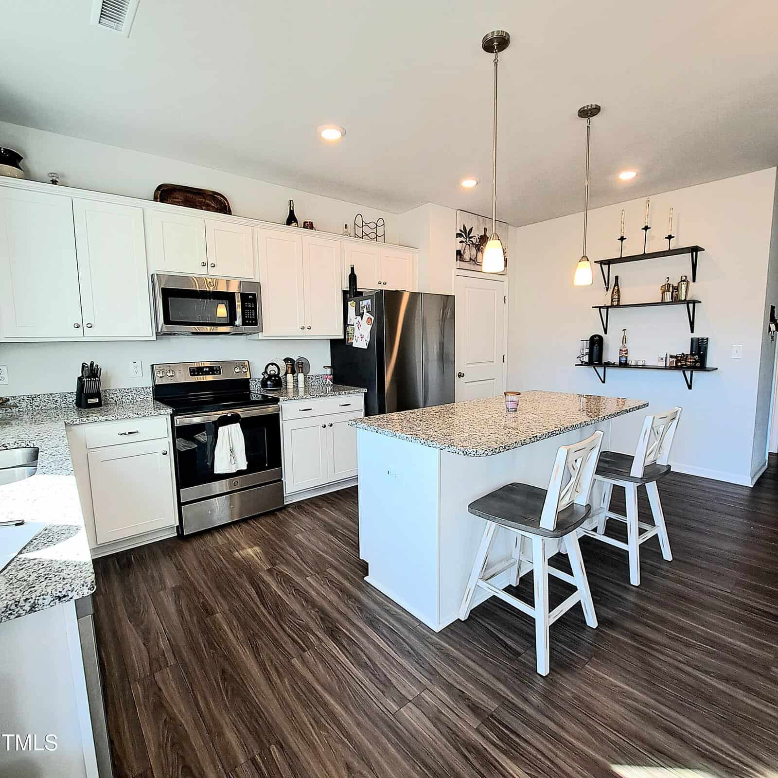 Modern kitchen with white cabinets, stainless steel appliances, a center island with two chairs, granite countertops, pendant lighting, and wooden floors. Open shelves with decor items are on the far wall.