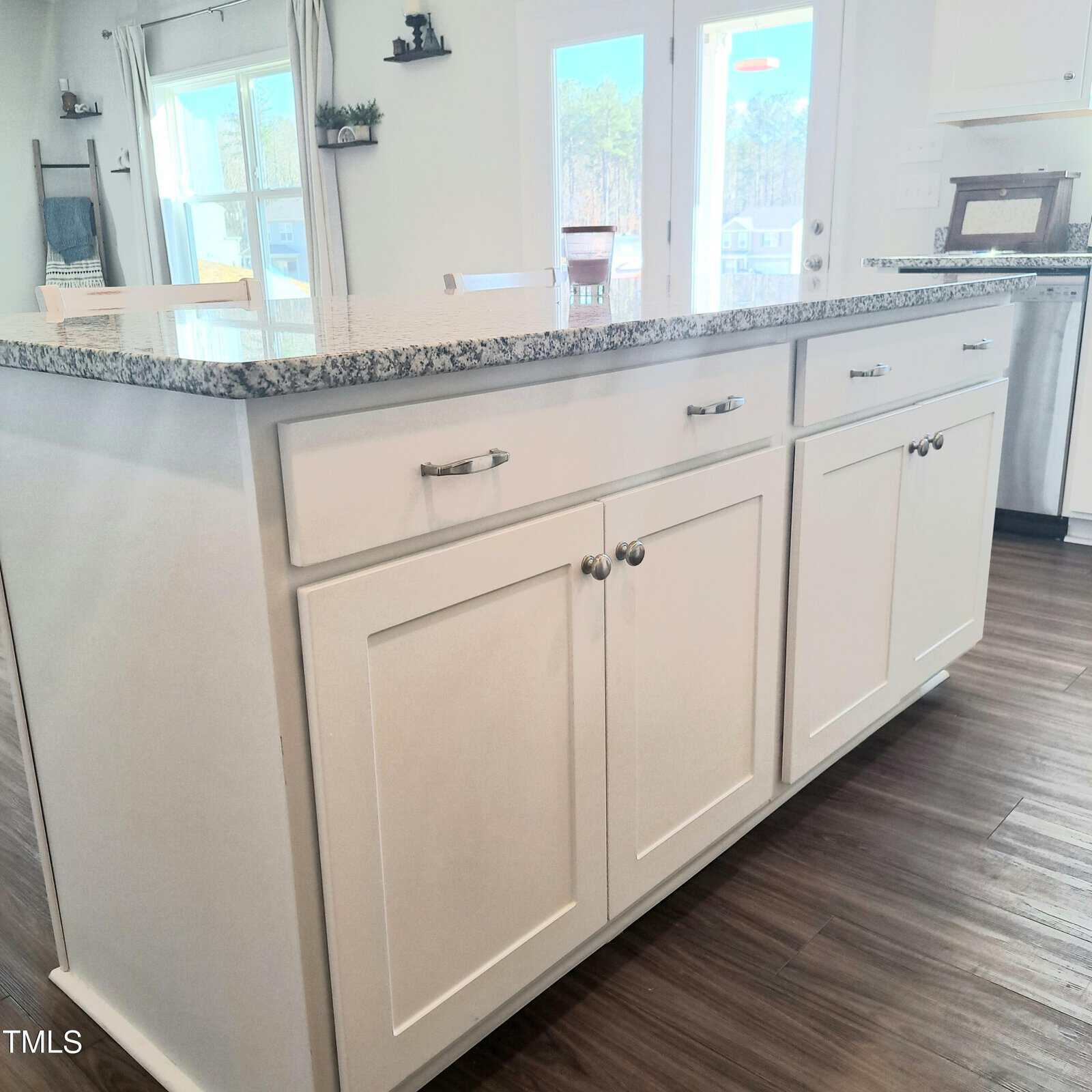A kitchen island with white cabinets and silver handles, topped with a gray granite countertop, stands on a wooden floor in a bright, modern kitchen with sliding glass doors.