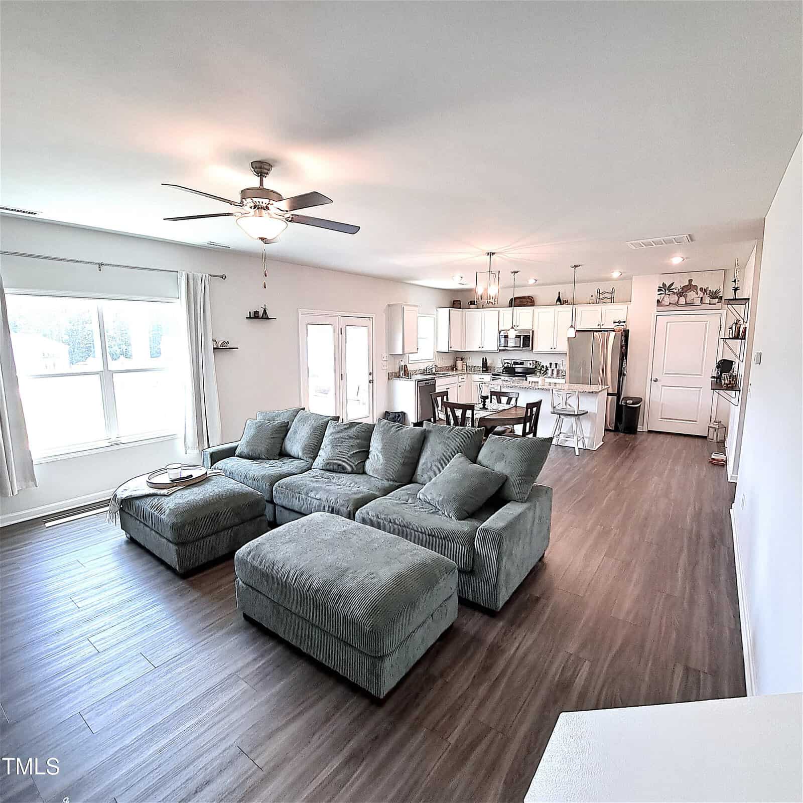 Open-concept living area with a large green sectional sofa and ottoman on wood flooring, adjacent to a kitchen with white cabinets and stainless steel appliances, and a dining table; ceiling fan above.