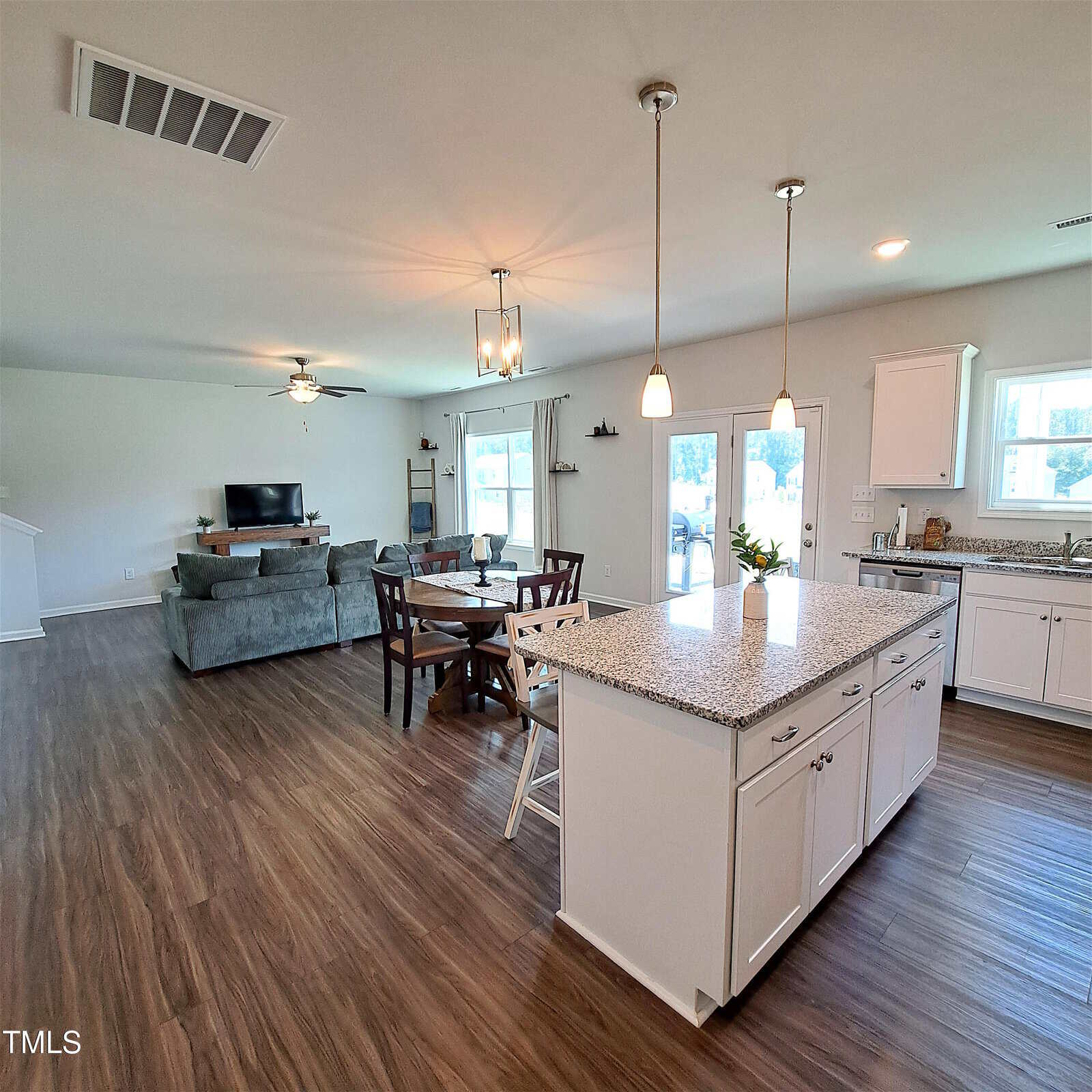 Open-concept living, dining, and kitchen area with wood flooring, white cabinets, granite countertops, and pendant lighting. There is a gray sofa, TV, round dining table, and glass doors leading outside.