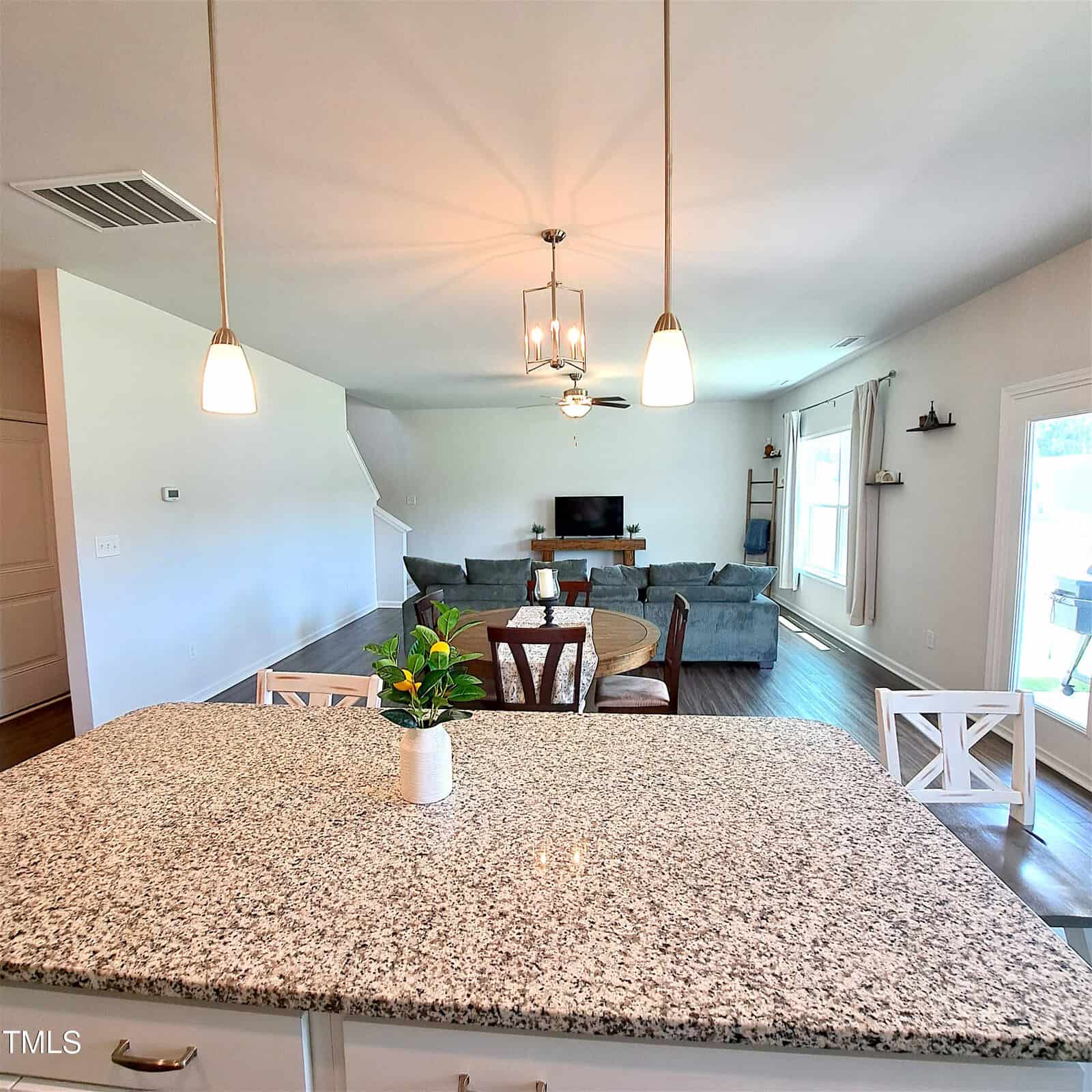 View from a granite kitchen island facing a living room with gray sofas, a round dining table, TV on a stand, wood flooring, large windows, and two pendant lights overhead. A small plant sits on the island.