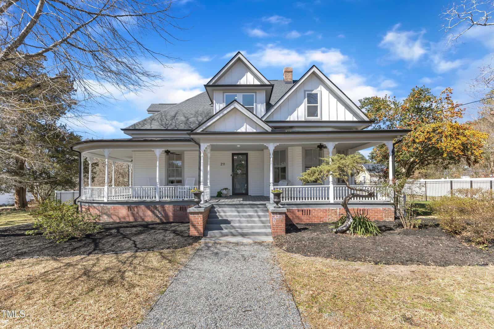 A large white two-story house with a wraparound porch, gabled roof, and brick steps leading to the front door. The yard is landscaped with mulch, grass, and a few trees.