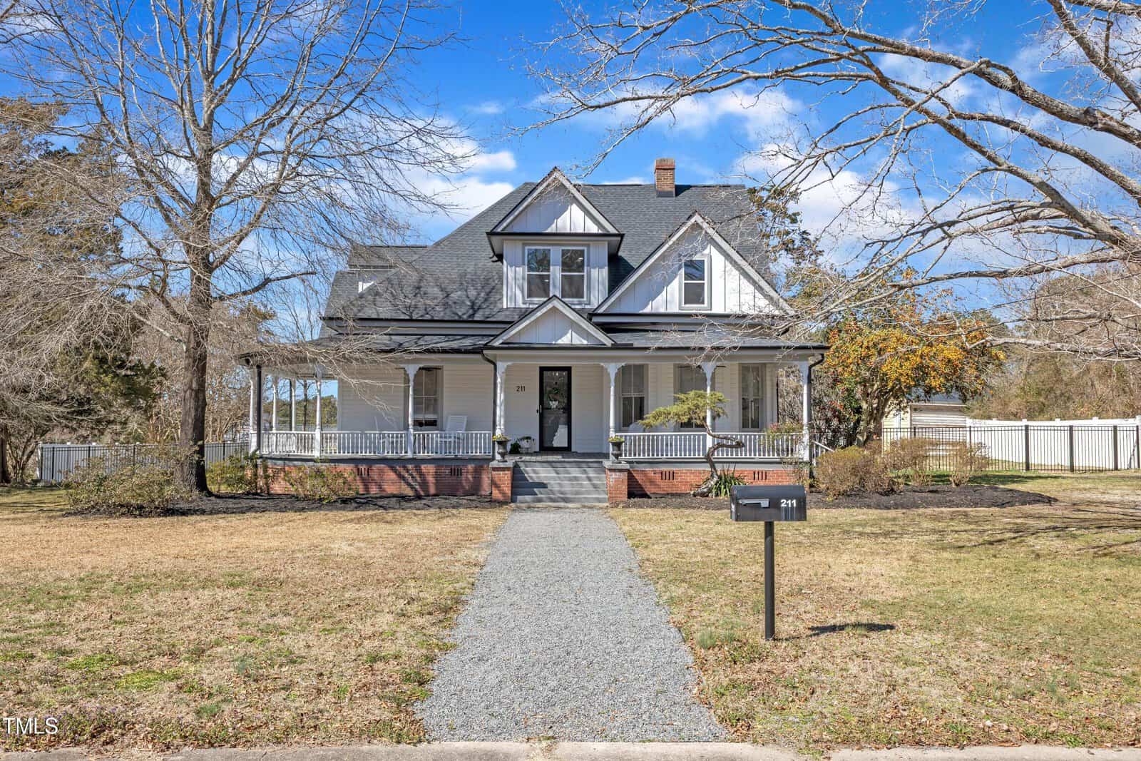 A two-story white house with a large front porch, brick foundation, and gabled roof sits behind a gravel walkway. The yard has leafless trees and a black mailbox labeled "211." The sky is blue with scattered clouds.