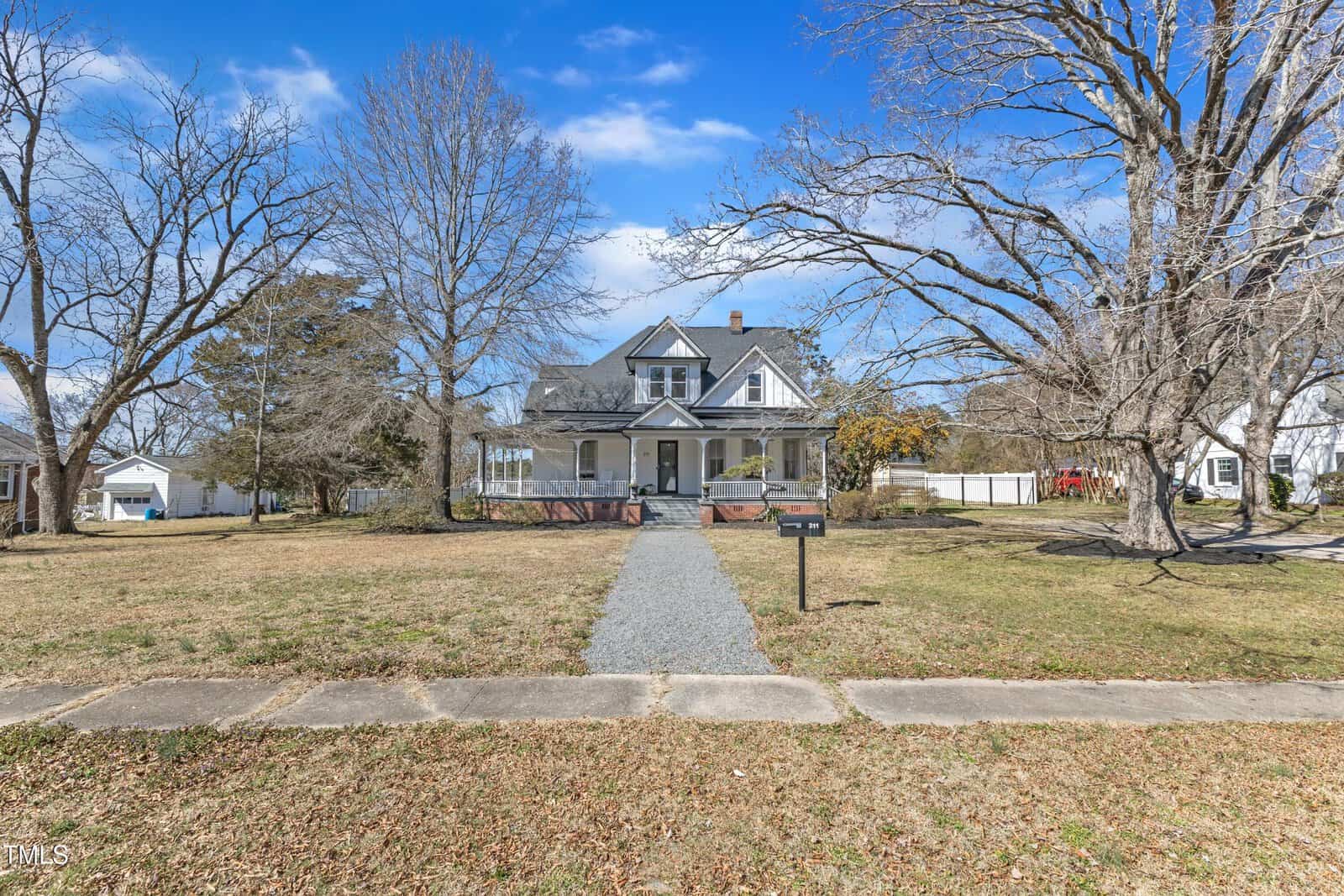 A two-story house with a large front porch sits at the end of a gravel walkway, surrounded by a spacious, leafless yard with mature trees under a blue sky. A mailbox stands near the curb.