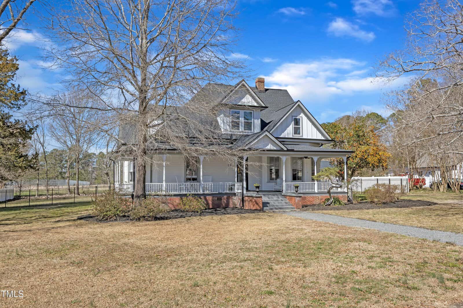 A large, two-story white house with a wraparound porch and gabled roof sits on a spacious yard with leafless trees and a curved gravel path leading to the front steps under a blue sky.