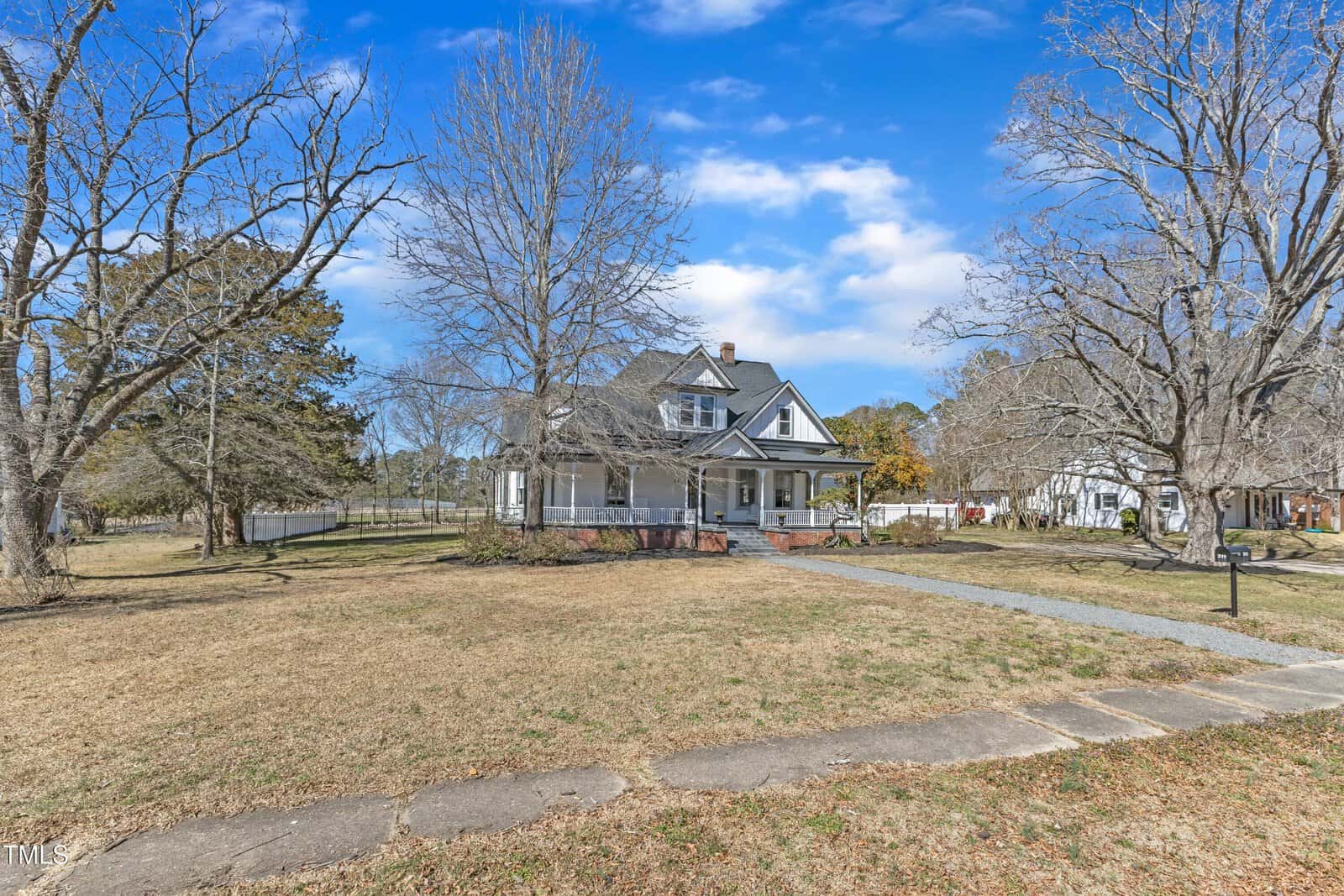 A large two-story house with a wraparound porch sits on a spacious, leafless yard with several tall trees and a brick walkway leading to the entrance. The sky is blue with scattered clouds.