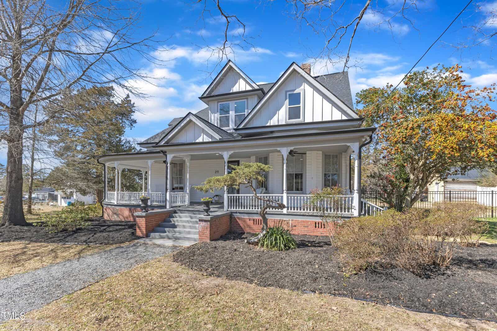 A two-story white house with a gabled roof, wraparound porch, gray steps, and brick foundation sits on a landscaped yard with bare trees and shrubs under a blue sky.