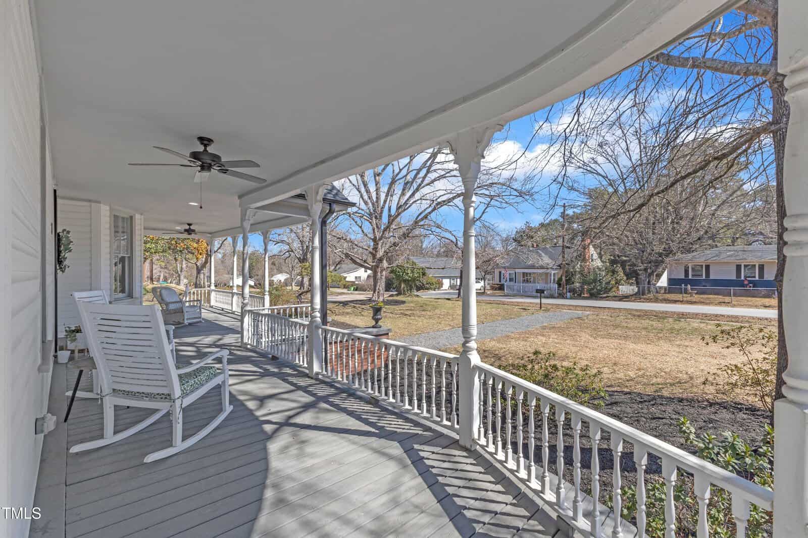 A wide, covered front porch with white railings, two white rocking chairs, ceiling fans, and a view of a grassy yard with trees and a paved driveway in a suburban neighborhood under a blue sky.