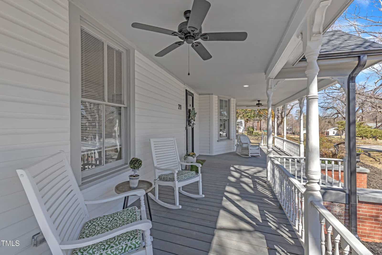 A covered porch with white railings and columns, gray wooden floor, white rocking chairs with green cushions, ceiling fans, and potted plants. Houses and trees are visible in the background.