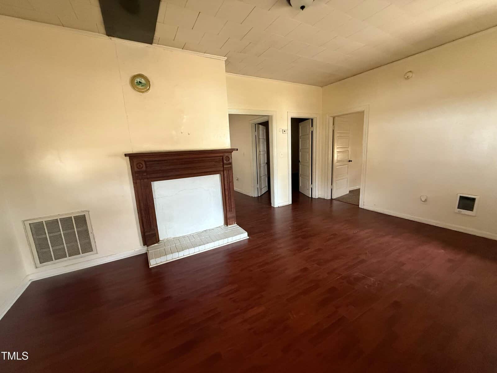 A room with reddish-brown wood flooring, cream-colored walls, a white fireplace with a wooden mantel, and multiple doorways leading to other areas. Ceiling panels and vents are visible.