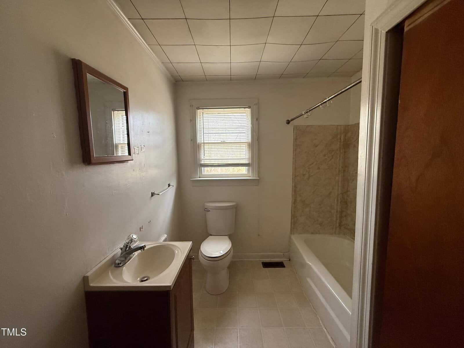 A small bathroom with a single-sink vanity, a wall-mounted mirror, a toilet, a window with blinds, and a bathtub with a shower curtain rod. The walls are light-colored and the floor is tiled.