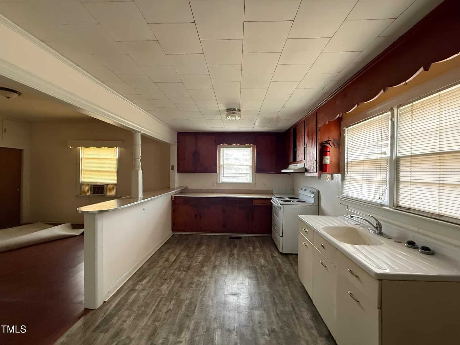 A kitchen with wood-paneled lower cabinets, white upper cabinets, white stove, and a white sink. The floor is vinyl, and there are two windows with blinds. The ceiling has square tiles, and an open doorway leads to another room.