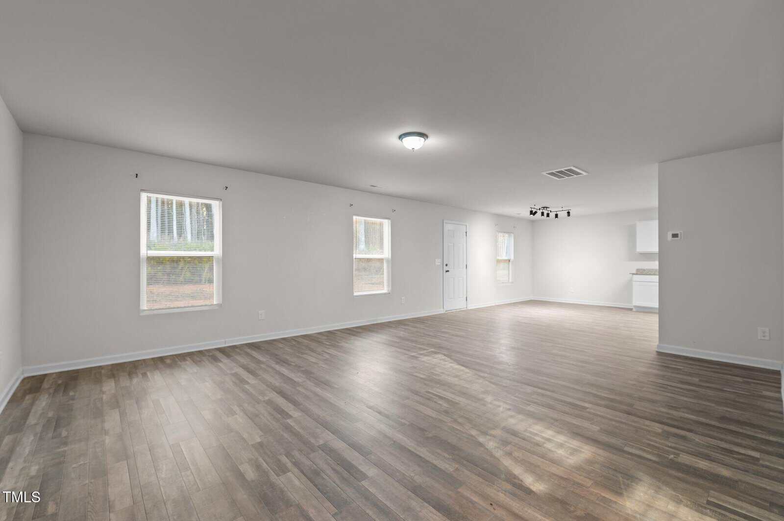 A spacious empty room with light gray walls and hardwood flooring. It has two windows on the left wall and two more at the far end. There's a ceiling light in the middle and a kitchen area visible to the back right.