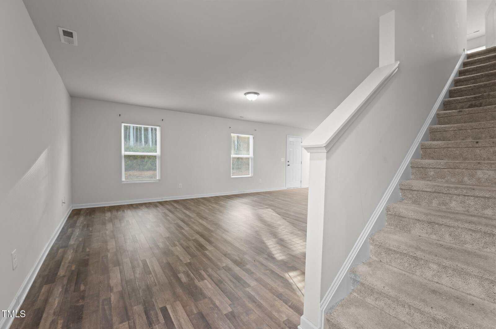 A vacant room with light-colored walls and wooden flooring. Two windows are on one wall, and a closed white door is visible. Carpeted stairs are in the foreground on the right, ascending to another level. A central ceiling light is lit.