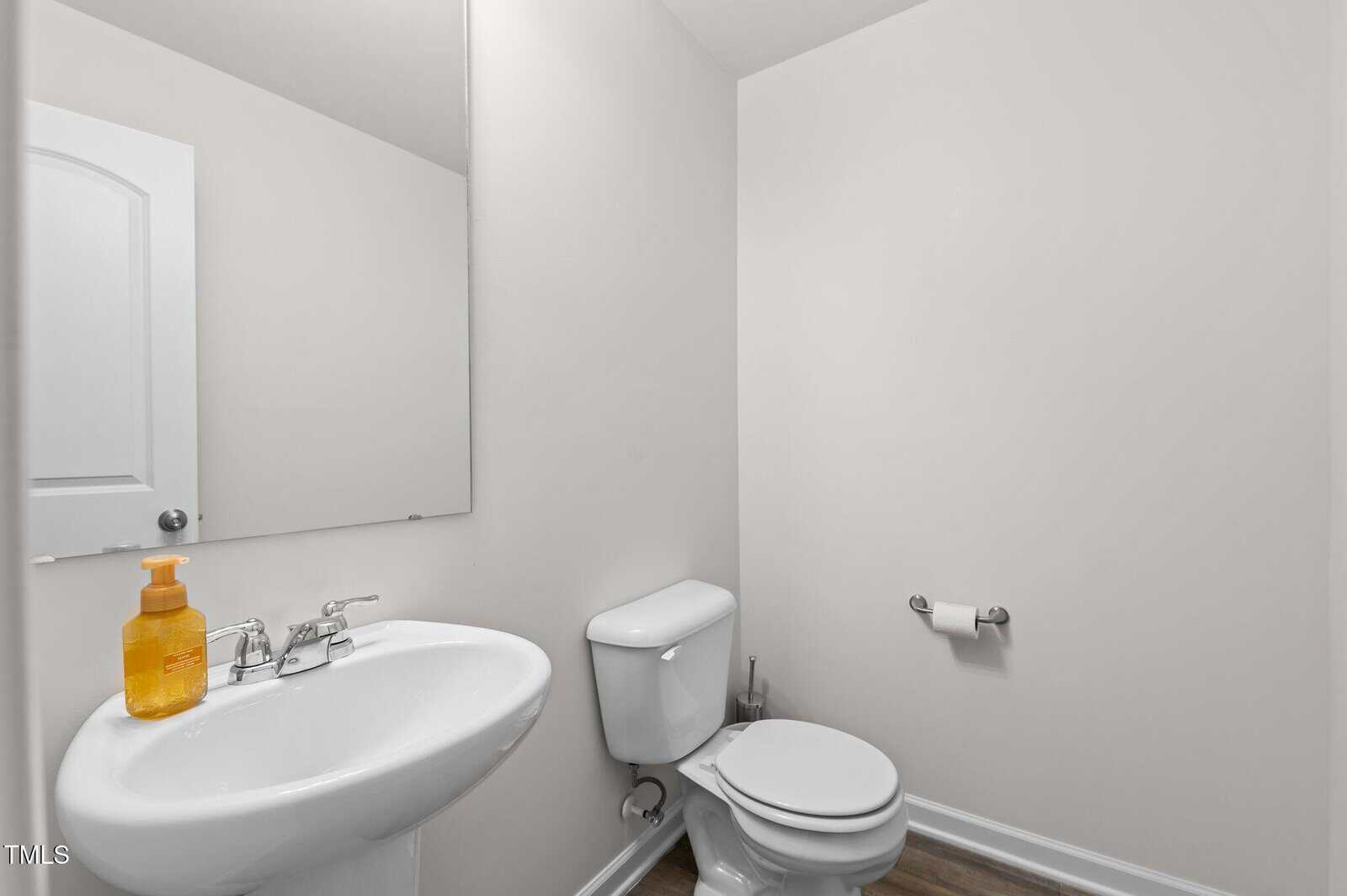 A small, white bathroom with a pedestal sink and a wall-mounted mirror above it. A bottle of orange soap is on the sink. There is a toilet next to the sink, and a toilet paper roll on a holder near the wall. The floor is wooden.