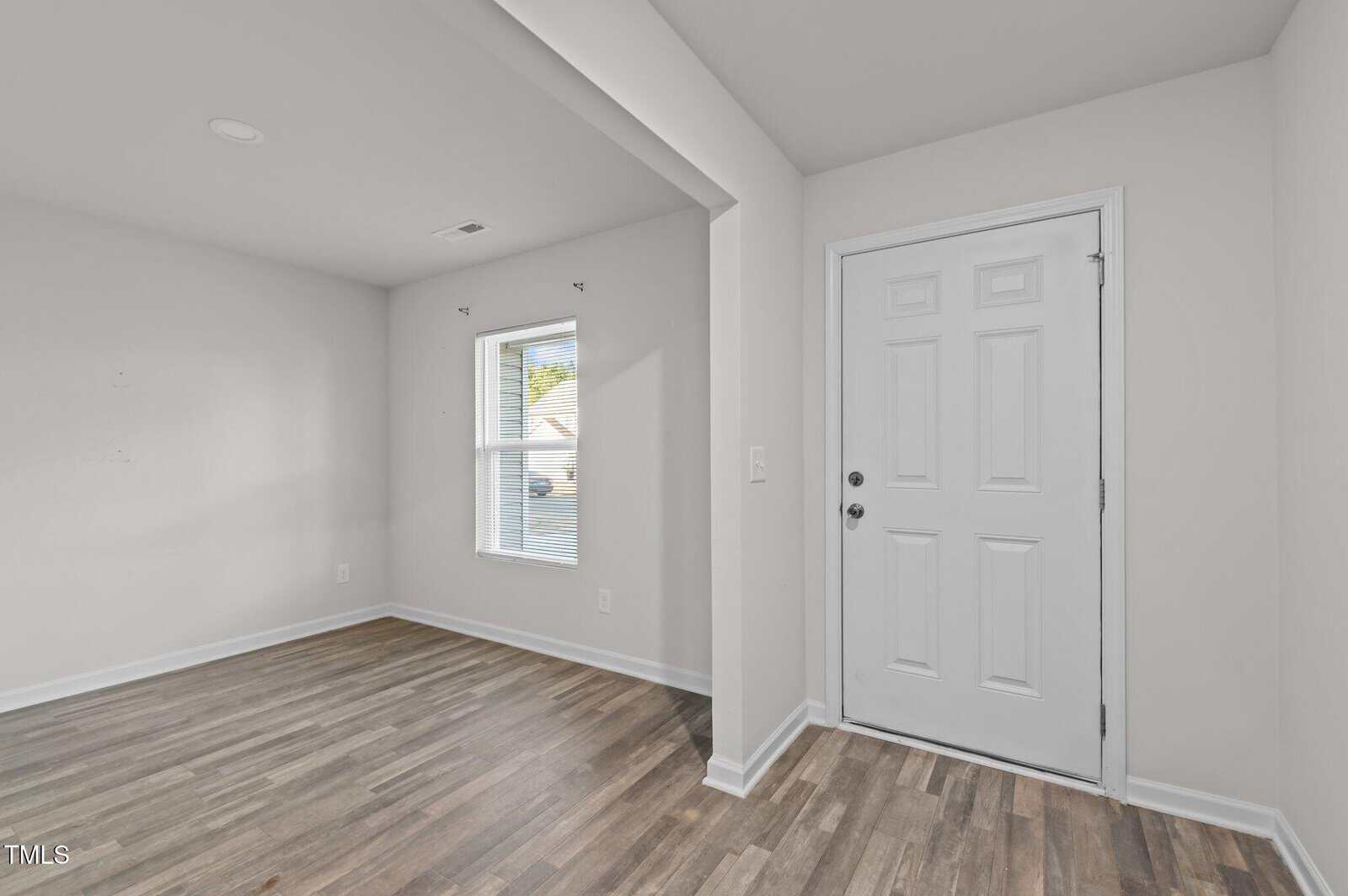 A small, empty room with light gray walls and wood-patterned laminate flooring. It features a window with blinds on the far wall and a white front door to the right. The space is well-lit with natural light.