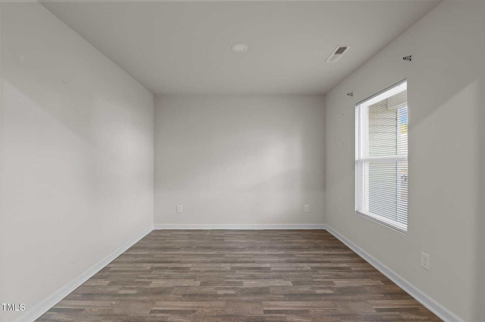 An empty, unfurnished room with light gray walls and a wooden floor. There is a window with blinds on the right wall, allowing natural light to enter. The ceiling has a light fixture, and the room appears clean and well-lit.