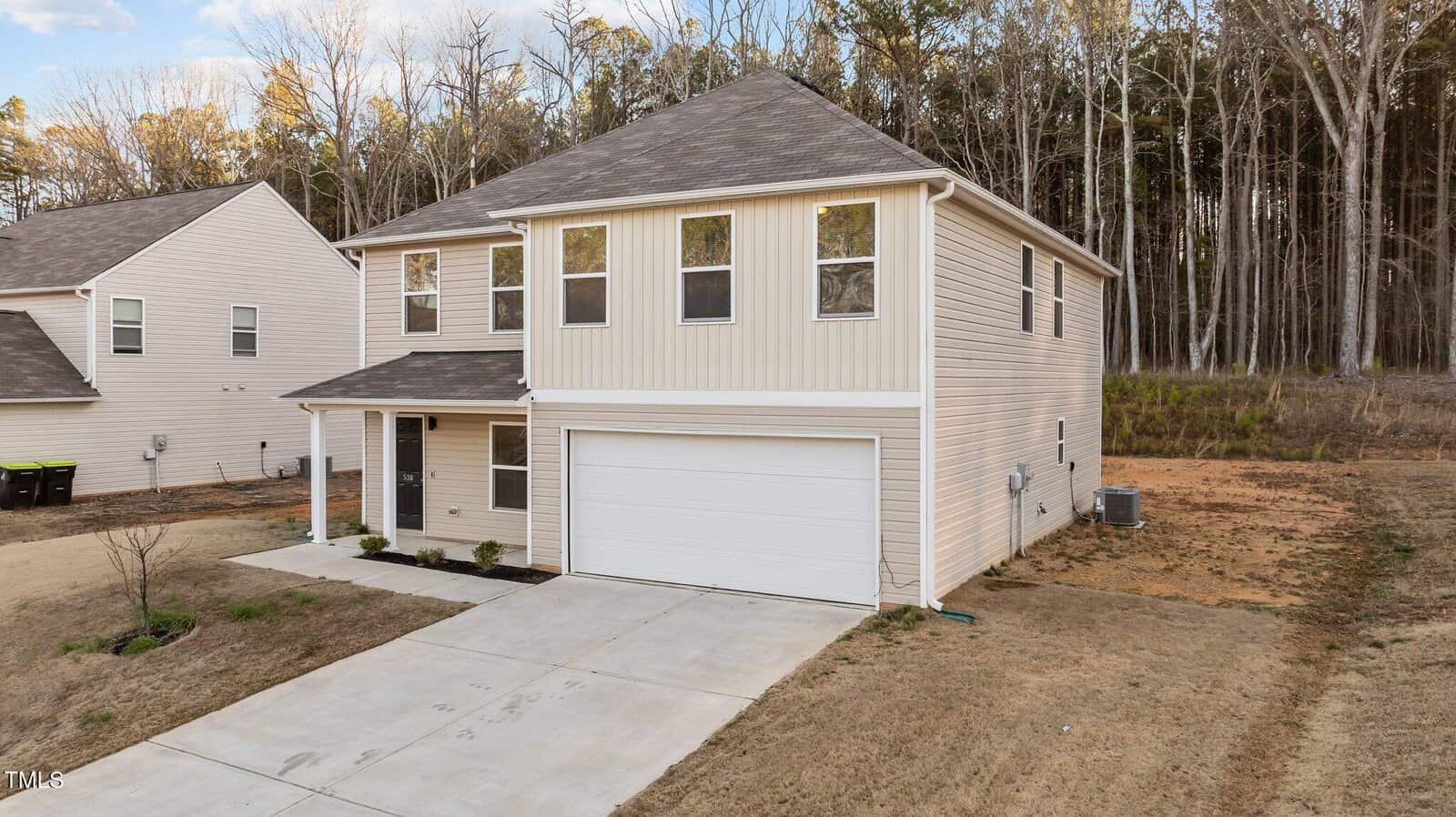 Two-story beige house with a two-car garage and a concrete driveway, situated on a newly developed lot. The house has a covered front porch and is surrounded by sparse grass and bare earth, with a forested area in the background.